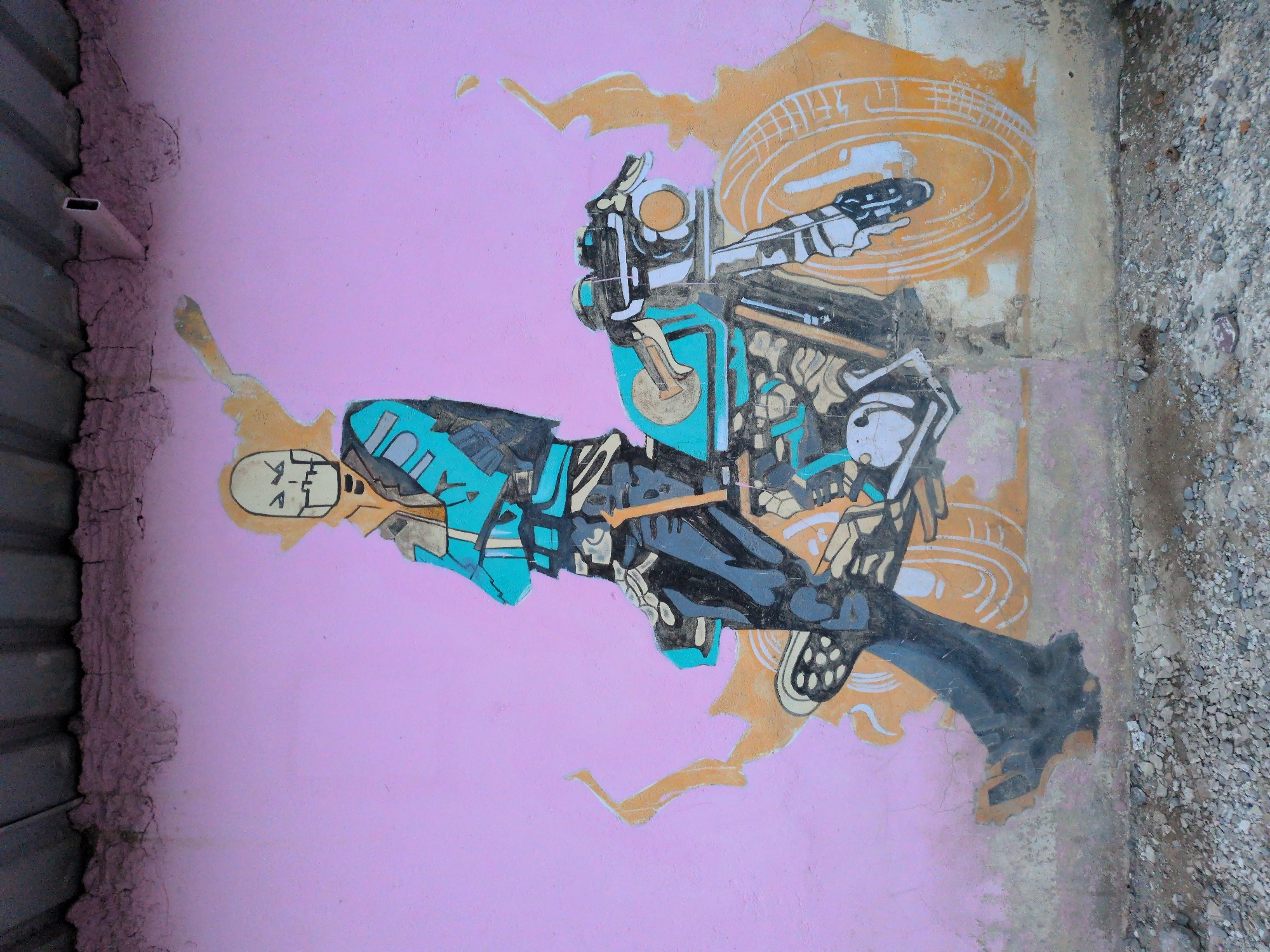 The image shows a mural painted on a pink, stucco wall in Leh, India. The mural depicts a skeleton-like figure wearing a teal jacket and dark pants, leaning against a stylized motorcycle. The figure has a skull for a head with a yellow hood. The motorcycle is predominantly teal and black with orange accents. Abstract orange shapes surround the figure and the motorcycle, possibly representing flames or energy. The wall appears weathered and cracked, with some areas of peeling paint. The bottom of the image shows the ground covered in gravel and some debris. Above the mural, a corrugated metal roof is visible.