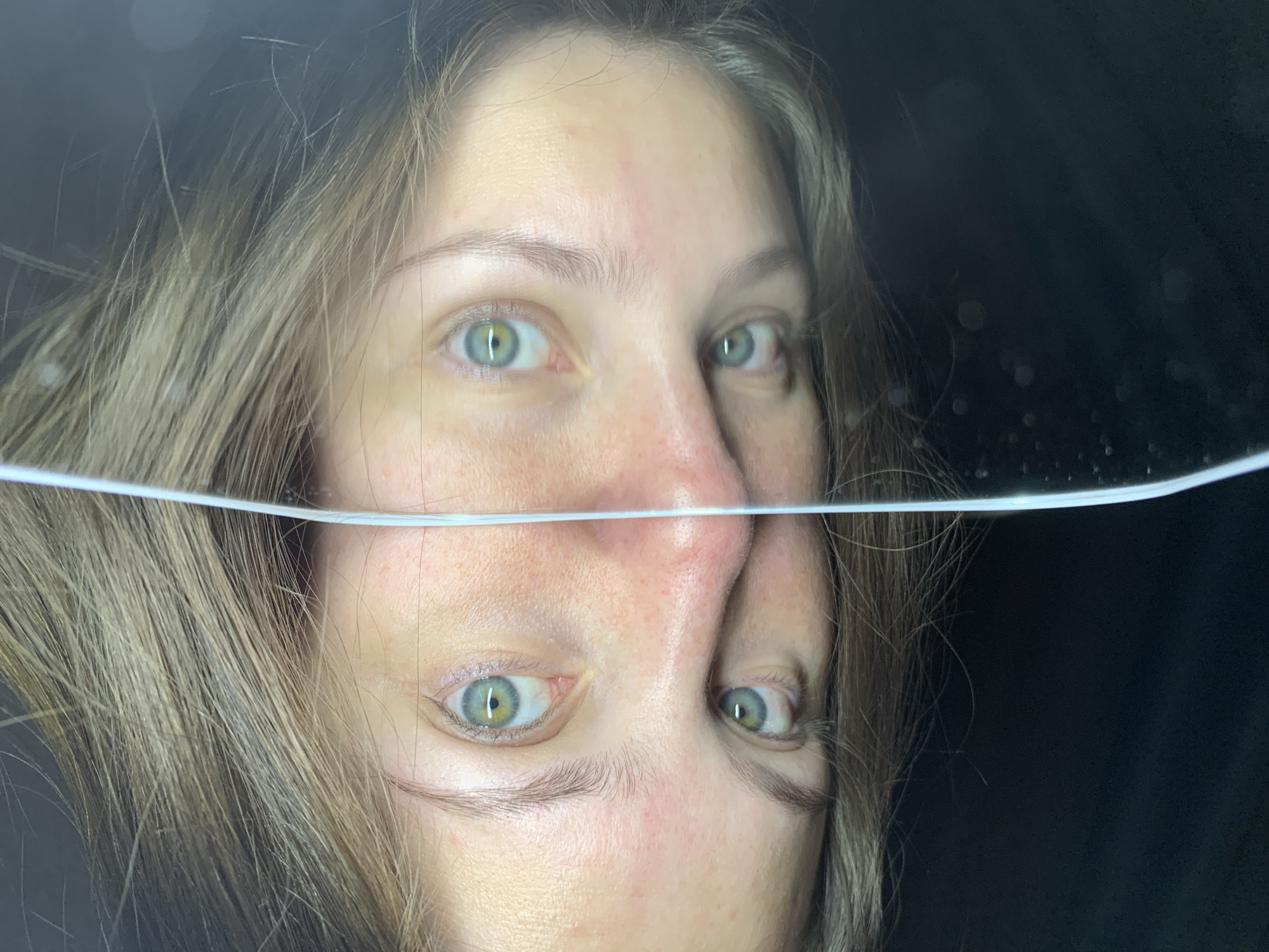 The image presents a close-up portrait of a person's face, uniquely bisected by a horizontal line, likely a water surface. The upper portion of the face, from the forehead to the bridge of the nose, is clearly visible above this line. The person has light brown or blonde hair framing their face and striking green or hazel eyes that are open and directly facing the viewer, conveying a neutral and contemplative expression.
Below the distinct horizontal line, the lower half of the face – including the nose, mouth, and chin – appears as an inverted reflection, suggesting the person is looking into or partially submerged in water. This reflection creates an optical effect where the reflected features are upside down relative to the upper part of the face. The reflected image is slightly distorted, consistent with being viewed through water.
The background behind the person is uniformly dark and indistinct, drawing full attention to the face and the intriguing visual division. No specific location cues, time of day, or weather details are discernible from the image itself. The overall composition suggests an artistic or experimental photograph exploring themes of perception and reflection. No visible text is present in the image. While the location context is Chaikovsky, Russia, the photograph lacks specific visual indicators of this geographical setting.