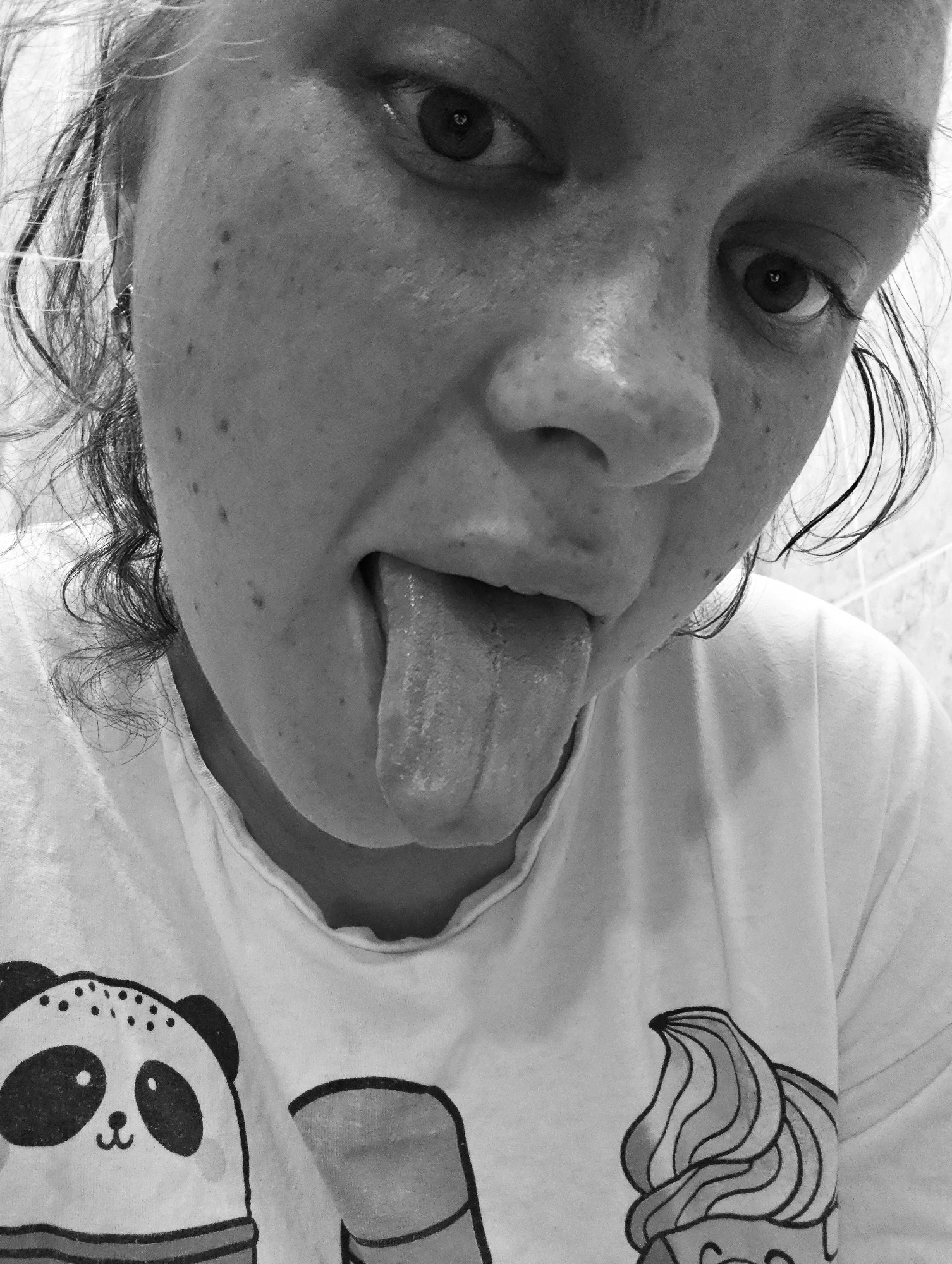 A close-up, black and white photograph depicts a person with their tongue extended. The person's face shows freckles and curly hair is visible on the left side of the frame. They are wearing a t-shirt with cartoon drawings, including a panda and a cupcake. The background appears to be a tiled wall. The image was taken in Chaikovsky, Russia.