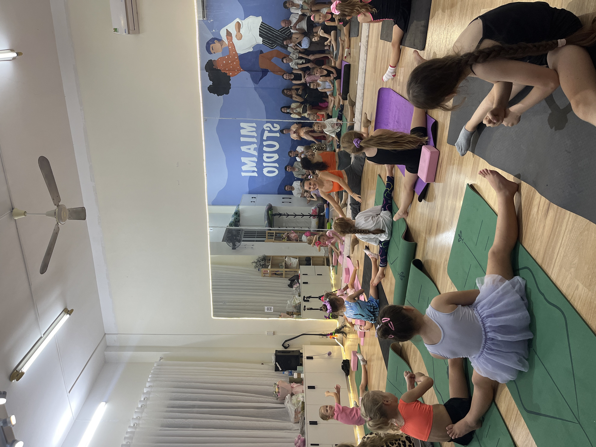 In a brightly lit room with a wooden floor, a group of young people are participating in a yoga class. The room features a large mural on the wall that reads "IMAIM STUDIO" in reverse, suggesting the image is a reflection. The mural depicts stylized figures in blue, white, and orange, with a person in the foreground wearing an orange polka-dot shirt and dark curly hair.
The participants are spread out on yoga mats of various colors, including green and purple. Some are in seated poses, while others are stretching or holding more advanced postures. Several children are present, some wearing colorful clothing and hair accessories, indicating it might be a children's yoga session. One young girl in the foreground is wearing a light purple tutu and black shorts.
In the background, shelves filled with various items and cabinets are visible. A large mirror reflects the scene, further enhancing the sense of space. The lighting appears to be a combination of overhead fluorescent lights and accent lighting along the top of the mural. The overall atmosphere seems energetic and engaged, with individuals focused on their practice. The presence of a ceiling fan suggests a warm environment, consistent with the tropical climate of Vietnam.