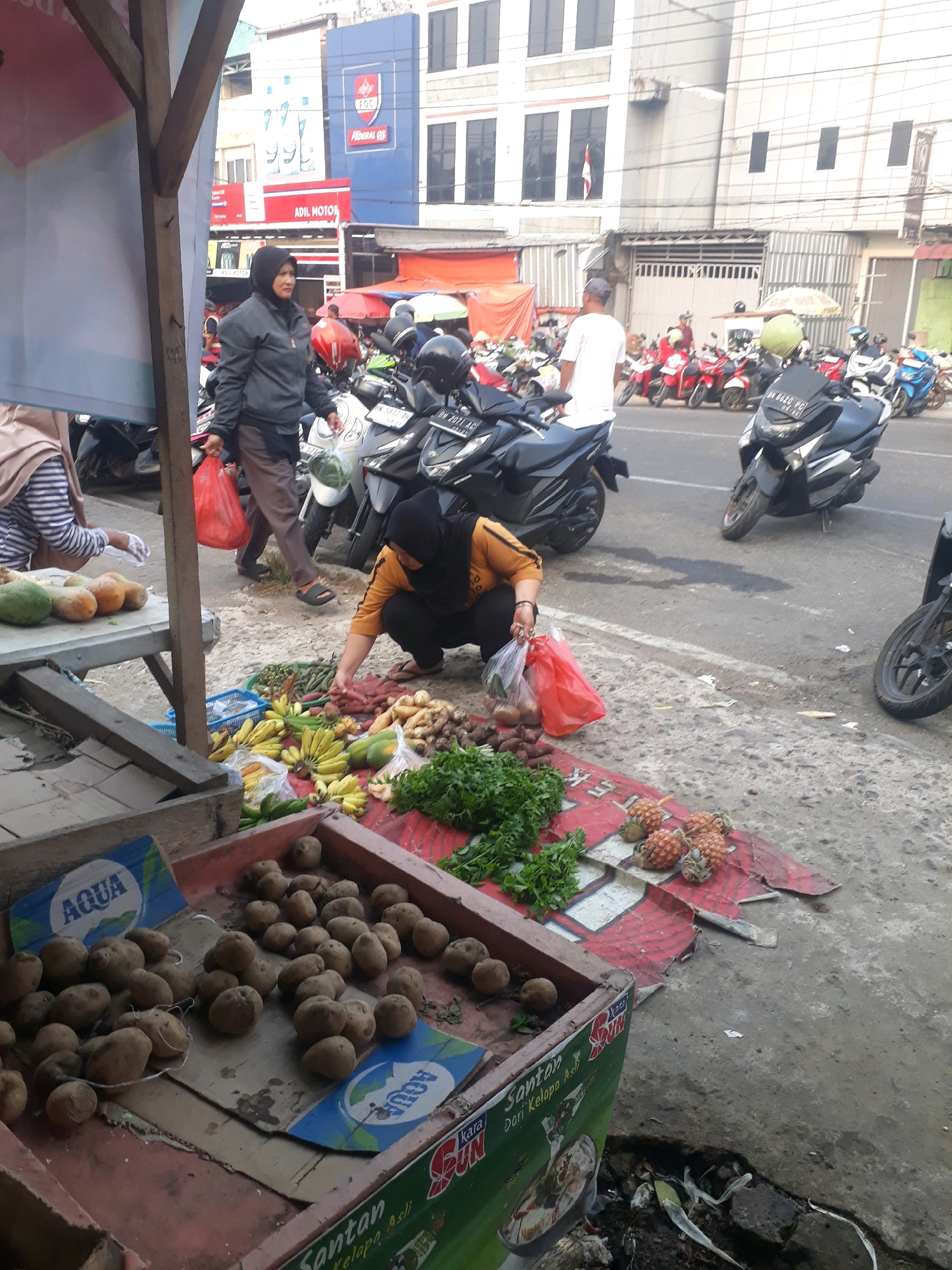 This is a street-level, medium-shot photograph taken in the city of Pangkalpinang, Indonesia. The scene depicts a busy marketplace with a variety of fresh produce displayed on the ground and in crates. In the foreground, a red plastic crate is filled with potatoes. To the right of the crate, a woman wearing a black hijab and an orange shirt is crouched down, sorting through vegetables and placing them into a red plastic bag. Next to her, a red and white patterned tarp is laid out on the ground, displaying pineapples and leafy green vegetables. In the background, there is a collection of motorcycles parked on the street, and several people are visible. Buildings with storefronts line the street, and a blue sign with "Federal Oil" is prominently displayed on one of the buildings. The overall atmosphere suggests a typical market day with commercial activity.