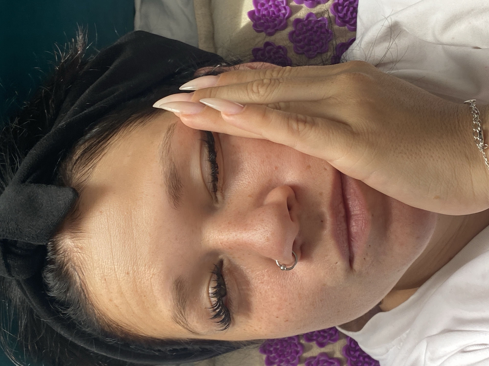 A woman with long dark hair, a black headband, and a white shirt is lying down. She has long artificial nails, a nose ring, and false eyelashes. Her right hand is pressed against her right cheek, and her eyes are closed. The background appears to be a teal wall and a light-colored surface covered with small purple flower-like objects, possibly an acupressure mat. The scene suggests a personal, relaxed setting, potentially a home environment in Yanino-1, Russia.