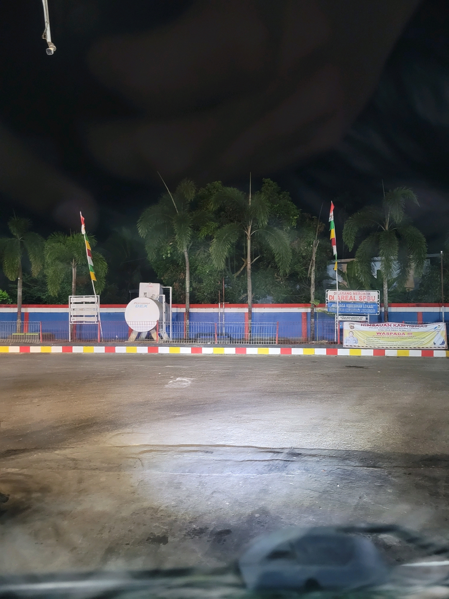 The image depicts an outdoor scene at night, likely in Seunebobaru, Indonesia. In the foreground, a paved surface with visible tire marks and reflective lighting from an unseen source dominates the lower half of the frame. A low wall with a red, blue, and white striped border runs horizontally across the middle ground. Behind the wall, several palm trees are visible, illuminated by artificial light. To the left of the image, a white structure resembling a price display for fuel is present. Further to the right, a large white cylindrical tank with the text "Pertamina DEX" is visible. A flagpole with an Indonesian flag is planted near the price display. A blue sign with white text stating "DILARANG MEROKOK DI AREAL SPBU" (No Smoking in the SPBU Area) and other regulations regarding environmental cleanliness is mounted on the wall. To the right of this sign, a yellow banner with text in Indonesian, including "HIMBAUAN KAMTIBMAS" (Public Security Appeal) and "WASPADA !!!" (Beware!!!), is displayed. The overall impression is that of a fuel station area at night, with some regulatory signage and a tanker. A security camera is visible in the upper left corner of the image, attached to a pole.