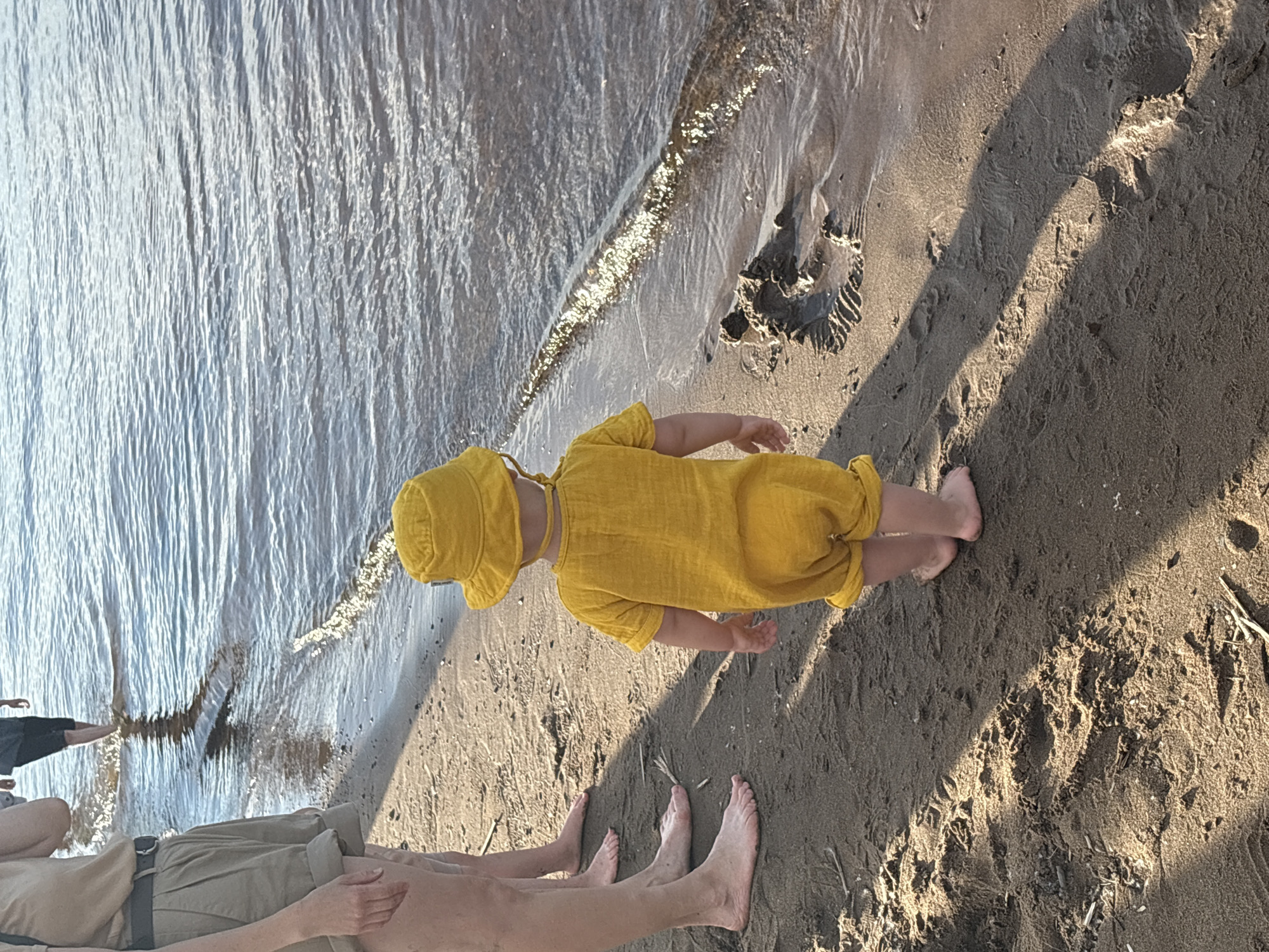 The image depicts a toddler wearing a yellow sun hat and a yellow jumpsuit standing on a sandy beach next to a body of water. The toddler is facing away from the camera and looking towards the water. In the background, the legs of at least two other people are visible, also standing on the beach. One person is wearing shorts and a belt, and the other is wearing dark shorts. The water is calm with ripples, and sunlight reflects off its surface. The beach sand shows evidence of footprints and some scattered debris. The scene appears to be a casual outdoor setting, possibly a park or recreational area by a river or lake. Chaikovsky, Russia, is known for its Kama River embankment, which is a popular spot for recreation and outdoor activities, making it a plausible location for this scene.
