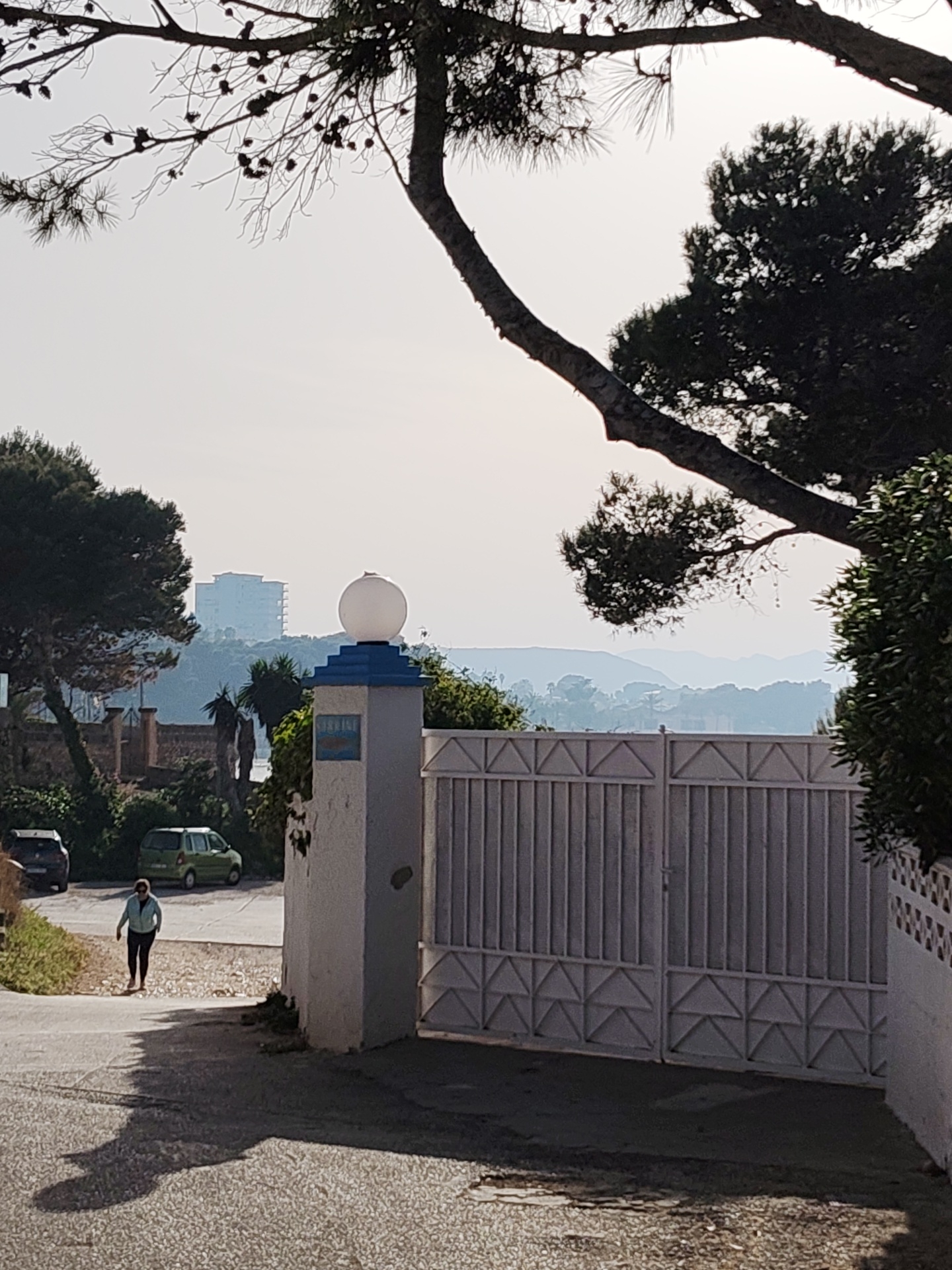 The image captures a street scene in Rotes, Spain, under a bright, overcast sky. A mature pine tree with visible cones dominates the upper portion of the frame, its branches arching over the scene. Below, a residential street slopes slightly downward. A woman in a light-colored jacket and dark pants walks along the left side of the street. Behind her are two parked vehicles: a dark-colored car closer to the camera and a green hatchback further away.
To the right, a white gate with vertical bars and a triangular pattern at the bottom is set into a white wall. A white pillar, topped with a spherical light fixture and a blue band, stands to the left of the gate. A sign on the pillar reads "MERIDINE" in blue letters.
In the background, a large, multi-story white building is visible on a distant hill. A hazy layer obscures the details of the coastline and mountains behind the building. Green vegetation lines the streets and surrounds the buildings, contributing to a coastal Mediterranean atmosphere.