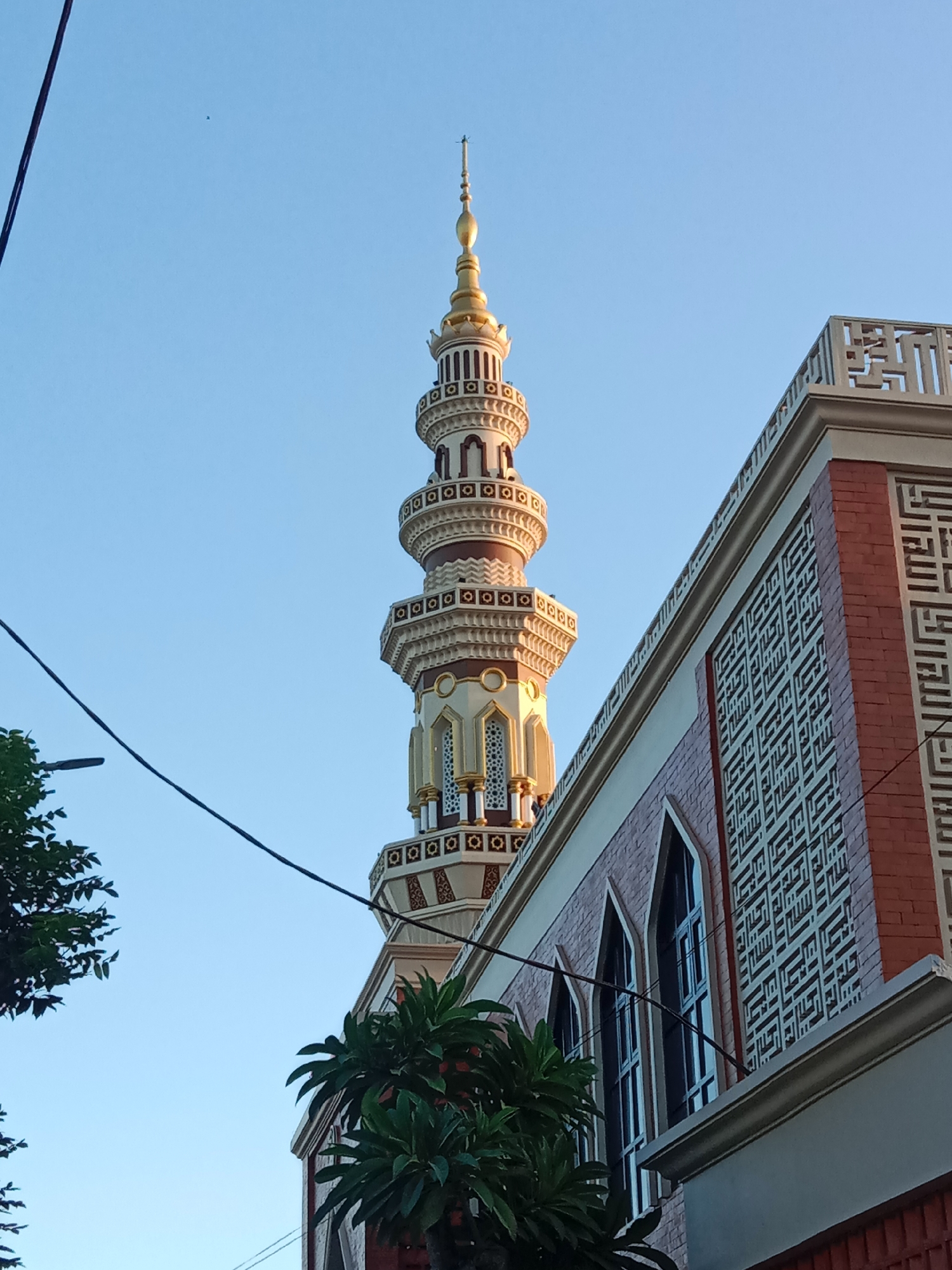 A minaret tower and a mosque are pictured under a clear blue sky in Ngingas, Indonesia. The minaret, a tall, slender structure with multiple levels, is topped with a golden spire. It features intricate decorative bands in white, gold, and dark red. Adjacent to the minaret, the mosque building showcases a facade of reddish-brown bricks adorned with white, geometric Arabic calligraphy patterns. Tall, arched windows with dark panes are visible on the mosque's side. Lush green foliage from trees partially obscures the lower parts of the scene. Overhead, dark electrical wires traverse the frame against the sky.