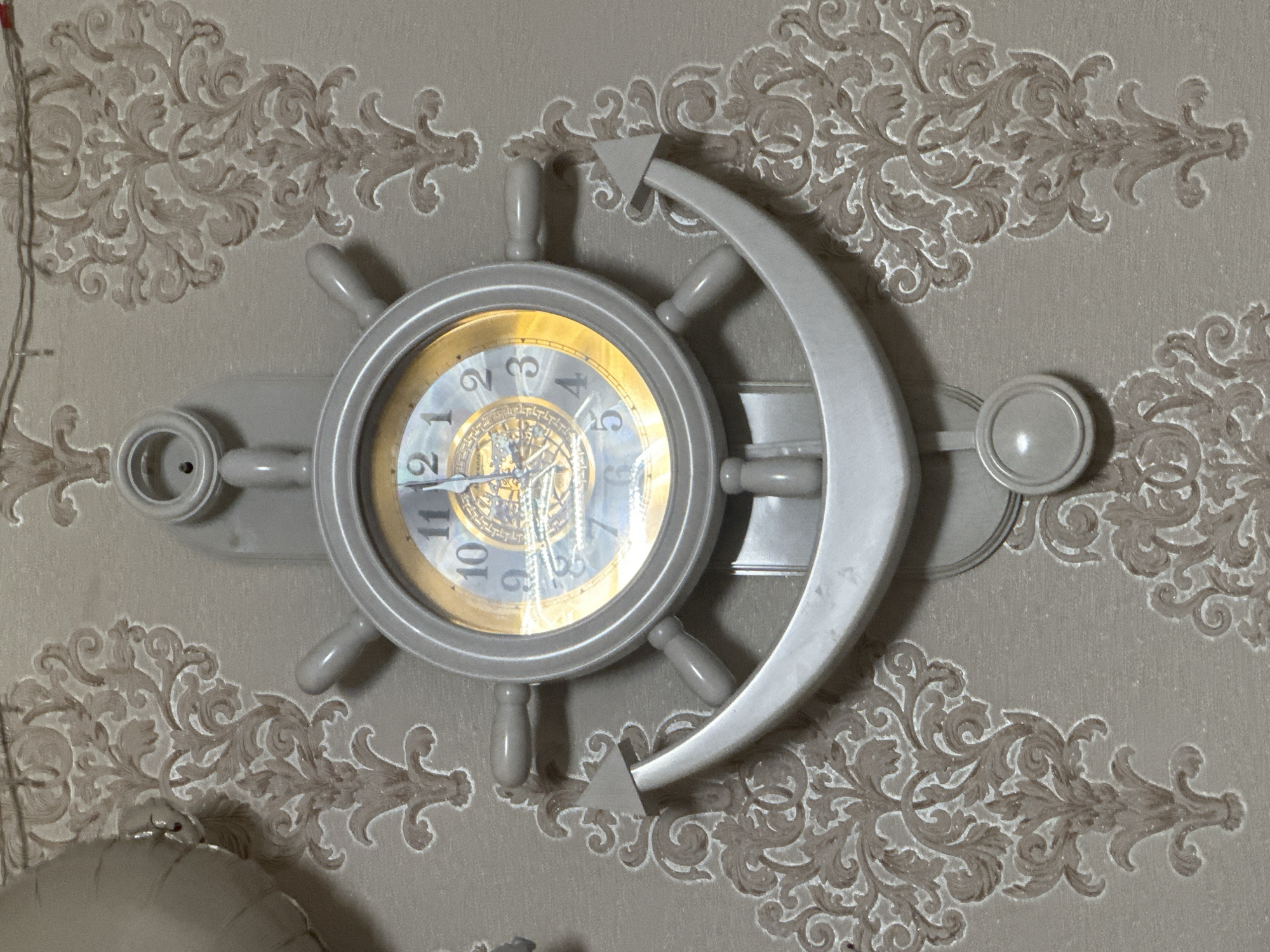 This is a close-up, horizontal shot of a decorative wall clock designed to resemble a ship's helm. The clock face is round with a gold-toned rim and a patterned background featuring what appears to be a compass rose. Arabic numerals mark the hours from 1 to 12, and the hands are visible, indicating a time around 10:55. The helm itself is a light gray, with a circular central hub and several spokes radiating outwards, each ending in a knob-like handle. A curved, crescent-shaped element extends from the right side of the helm. The clock is mounted on a wall covered in textured wallpaper featuring a repeating ornate, damask-like pattern in shades of beige and brown. The lighting is soft, suggesting an indoor setting. No people or specific weather conditions are visible. The location context provided is "Загородный, Russia," implying this is likely a residential or decorative setting within that area. There is no visible text on the clock face or surrounding elements, apart from the numerals on the clock.