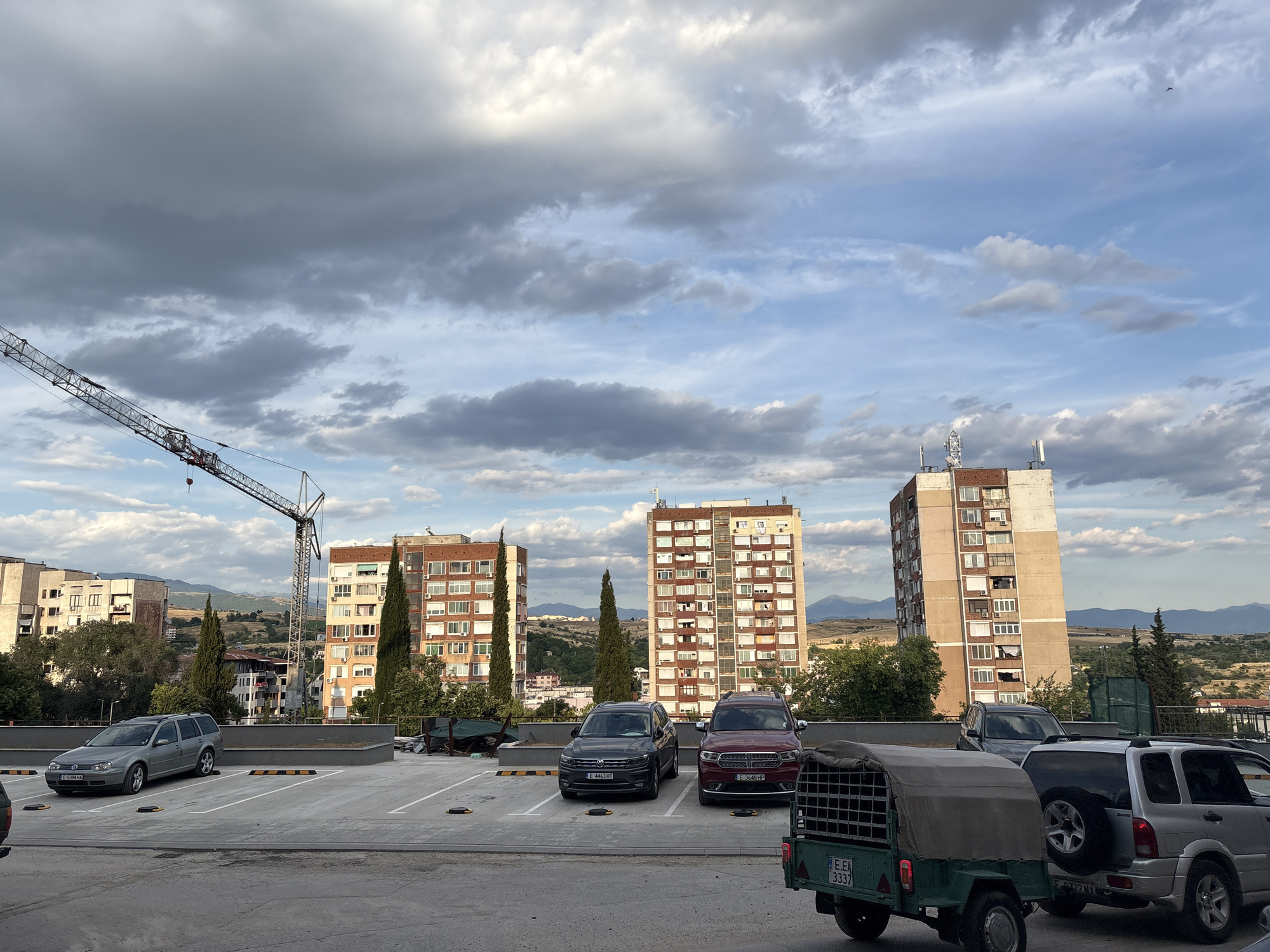 The image captures a scene in Sandanski, Bulgaria, featuring a rooftop parking lot with several vehicles. In the background, three tall apartment buildings dominate the skyline, with a construction crane positioned between two of them. The sky above is partly cloudy with a mix of gray and white clouds against a blue backdrop.
In the foreground, a gray Volkswagen station wagon is parked in the left section of the lot. Further back, a dark gray SUV and a maroon SUV are parked side-by-side. To the right, a silver SUV is visible, and a small green utility trailer is attached to the rear of a vehicle partially obscured from view.
The apartment buildings exhibit a mix of architectural styles, with balconies and numerous windows. The surrounding landscape appears somewhat hilly, with sparse vegetation and more distant structures. The overall impression is an urban environment with ongoing construction and residential areas under a cloudy sky.