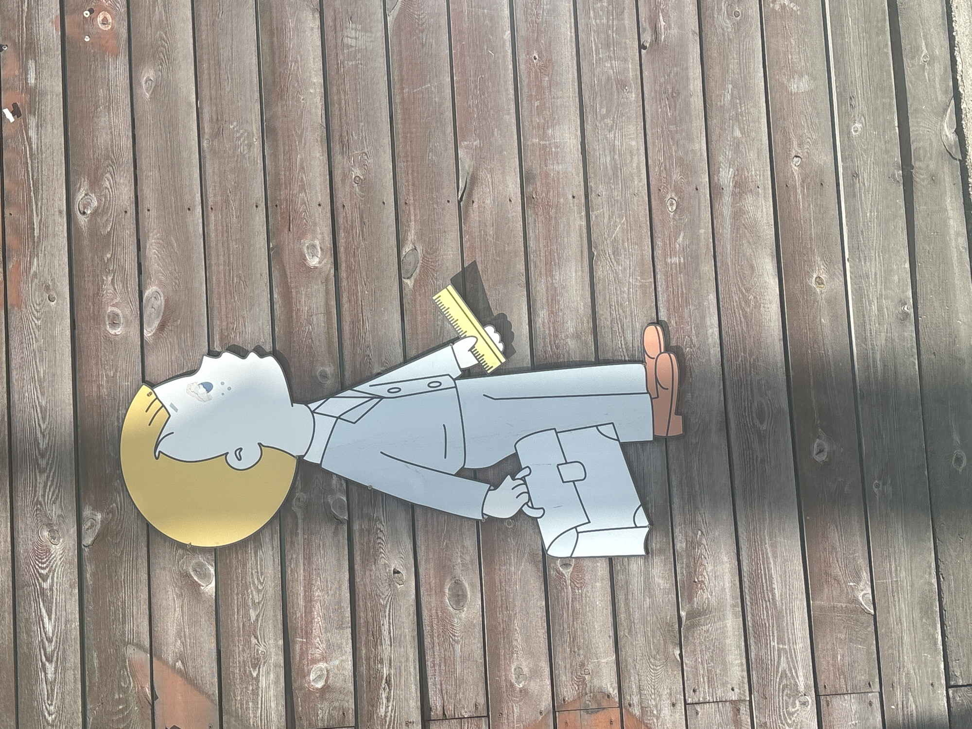 The image shows a cutout of a cartoon boy standing on a wooden wall. The boy is depicted in profile, facing right, with blond hair, blue eyes, and freckles. He is wearing a light blue suit and carrying a light blue briefcase in his left hand and a yellow ruler in his right hand. The wooden wall is made of horizontal planks. The image is taken in Novosibirsk, Russia.