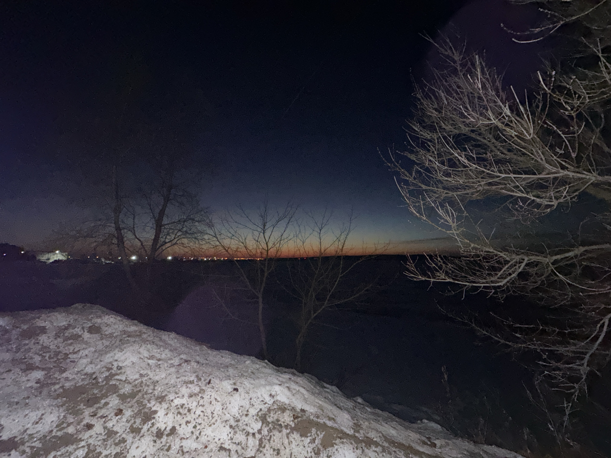 The image depicts a dark, winter scene in Chaikovsky, Russia. In the foreground is a snow-covered mound. Behind the mound are bare trees silhouetted against a dark sky, with a faint orange glow visible on the horizon, likely from city lights. There are more lights in the distance to the left and middle of the horizon, which could indicate the city center. The sky is a deep gradient of blue and dark grey. The scene appears to be a natural area on the edge of the city. No people or animals are visible.