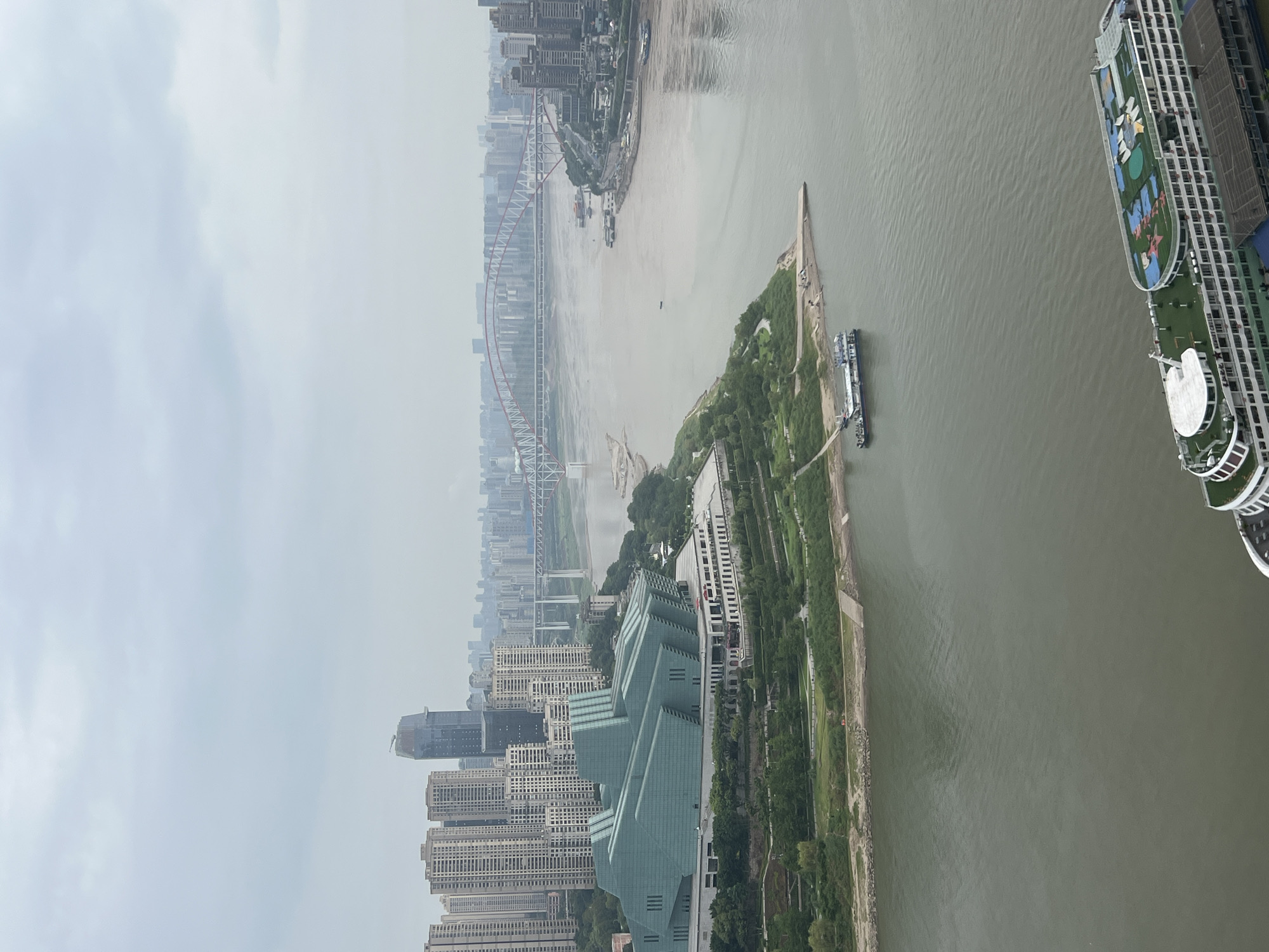 The image shows a wide, calm river flowing through a dense urban landscape under a hazy sky. The foreground features a large, multi-deck passenger ship with green and white detailing, its top deck covered in what appears to be artificial turf and recreational equipment. In the mid-ground, a distinctive, geometrically shaped modern building with a greenish-blue glass facade stands prominently on the riverbank, surrounded by green parkland. Beyond this building, a vast river stretches into the distance, spanned by multiple bridges, the most striking of which is a large red and white arch bridge. The background is dominated by a sprawling cityscape of numerous high-rise buildings and towers, indicative of a major metropolitan area. The overall impression is one of a modern city integrated with its natural waterways. The location is Yuzhong District, China.