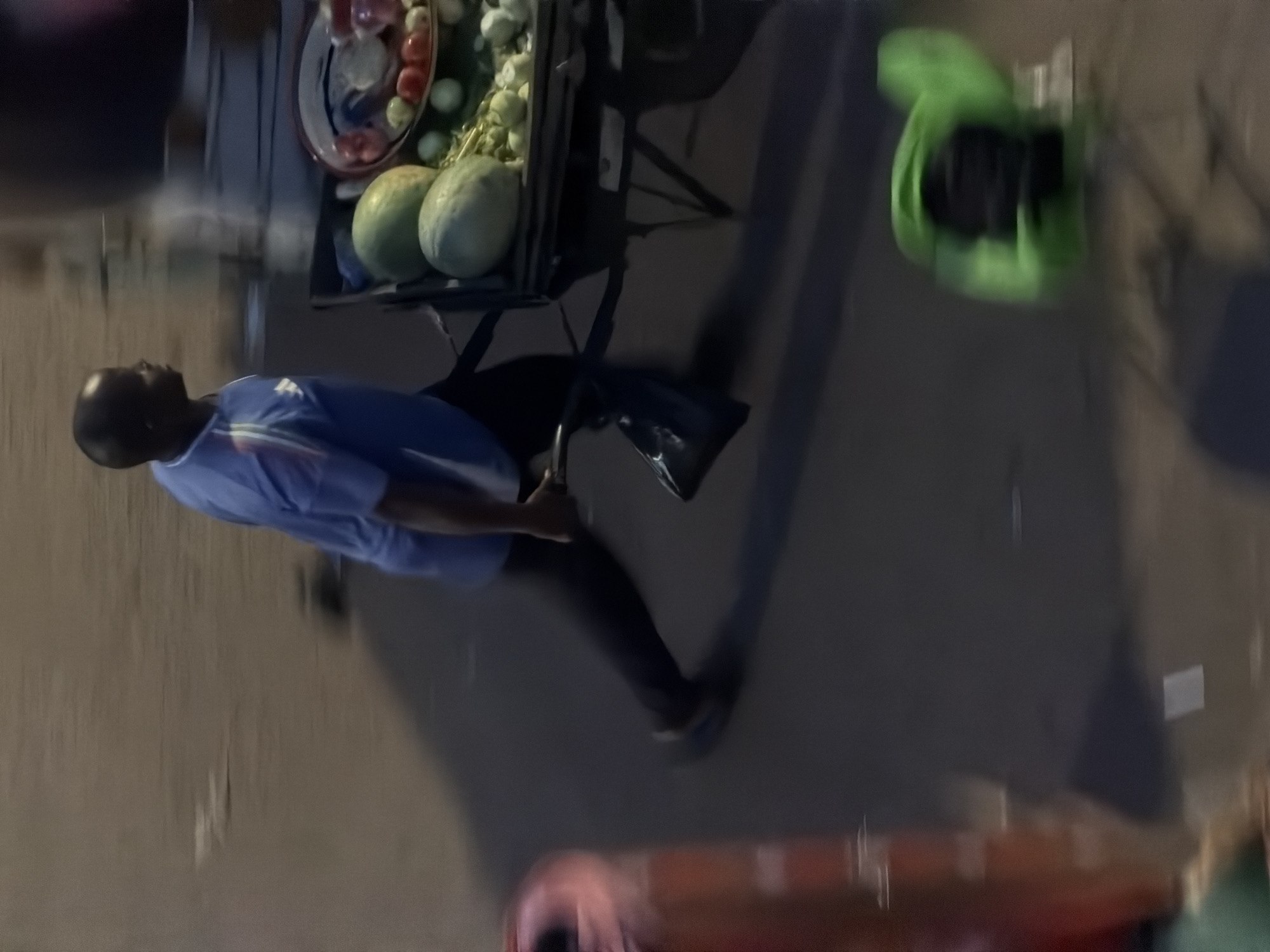 In Bukuru, Nigeria, a man in a blue shirt and dark pants pushes a wheelbarrow filled with fruits, including watermelons and what appear to be tomatoes and other vegetables. The wheelbarrow has a black frame and a woven basket. The man's face is partially obscured as he walks, his body in motion. To the left, a blurred orange object, possibly a traffic cone or barrel, is visible. In the background, the environment is indistinct due to motion blur and low light, suggesting an outdoor setting, likely a street or market area. A bright green object, perhaps a bag or tarp, is on the ground to the right, also blurred. The overall scene captures a moment of commerce or transport in a bustling urban environment.