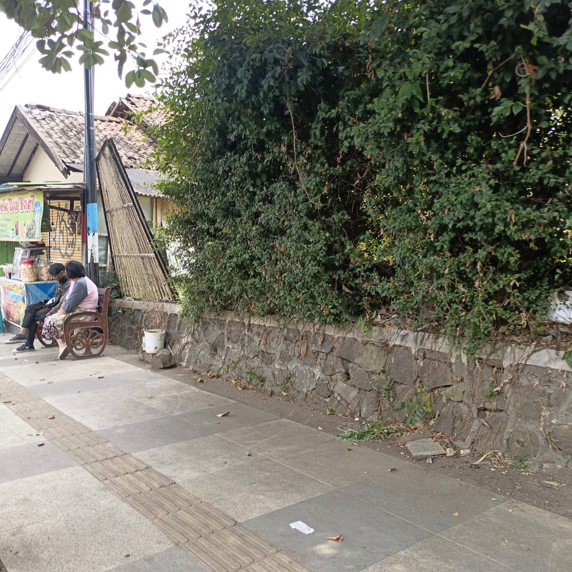 This image captures a scene on a sidewalk in Salatiga, Indonesia, adjacent to a street vendor's stall and a lush green wall of foliage. Two individuals are seated on a decorative bench, facing away from the camera. The person on the left is wearing a dark jacket and pants, while the person on the right is in a pink and grey long-sleeved shirt and a patterned skirt. The vendor's stall, partially visible on the left, has a banner with Indonesian text and appears to be selling drinks and snacks. A metal pole with blue and red accents stands between the stall and the bench. The sidewalk itself is paved with grey tiles, featuring a tactile paving strip. Behind the bench, a stone wall separates the sidewalk from the dense greenery. A white plastic bin sits on the ground near the wall. The environment suggests a public outdoor space, likely a commercial or pedestrian area.