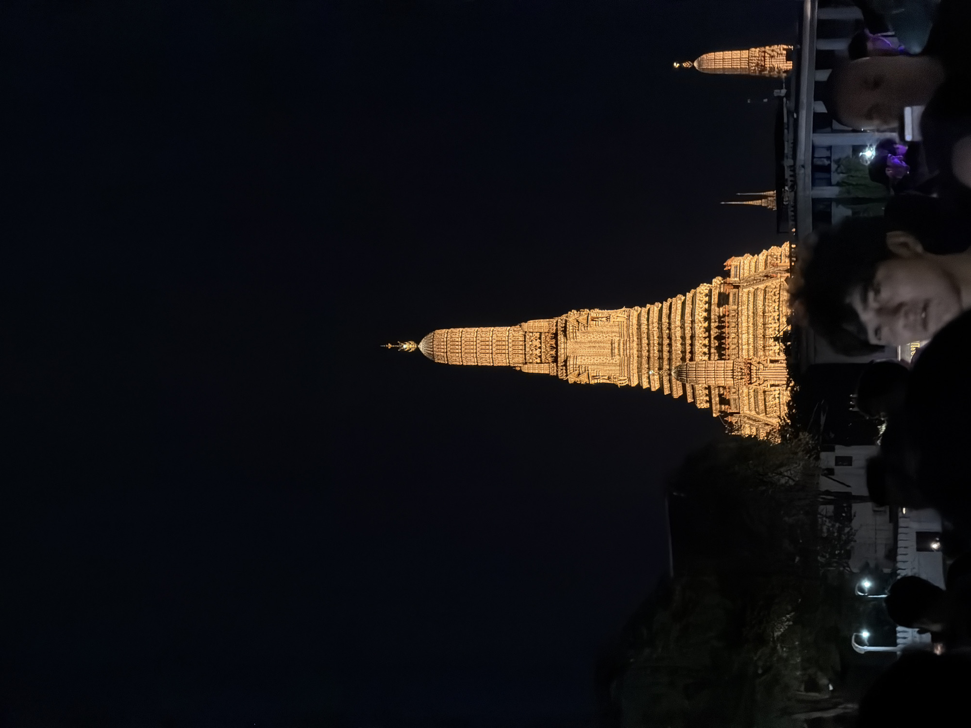 An evening shot captures the iconic Wat Arun temple, illuminated by warm lights, against a deep black night sky. The temple's tiered structure, adorned with intricate details, dominates the upper center of the frame. In the foreground, the silhouettes of several people are visible, their faces mostly obscured by shadow, suggesting they are observing the illuminated temple. The people appear to be standing outdoors, possibly on a promenade or viewing area, with hints of surrounding trees and buildings in the lower left and right portions of the image. The overall atmosphere is one of nighttime tourism and cultural appreciation in Bangkok, Thailand.