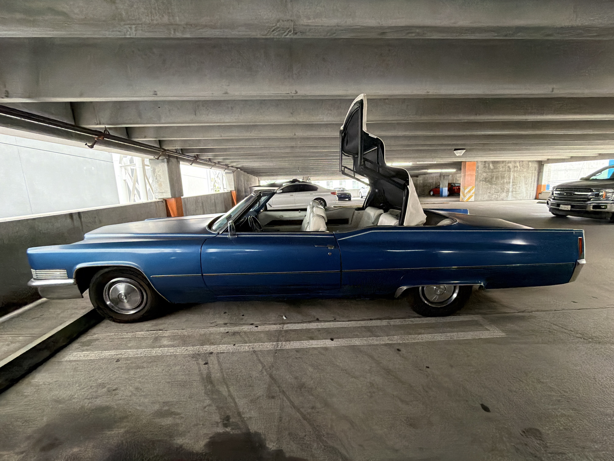 A blue 1960s Cadillac Eldorado convertible with its top partially raised is parked in an open-air parking garage in Los Angeles, California. The car is positioned diagonally in a parking spot, facing towards the right side of the frame. The interior of the car appears to be light-colored leather. In the background, other vehicles are visible, including a white sedan and a dark-colored pickup truck. The parking garage structure is made of concrete, with visible beams and supports overhead. Natural light filters down from the open ceiling. The overall impression is of a classic car displayed in a modern urban setting.