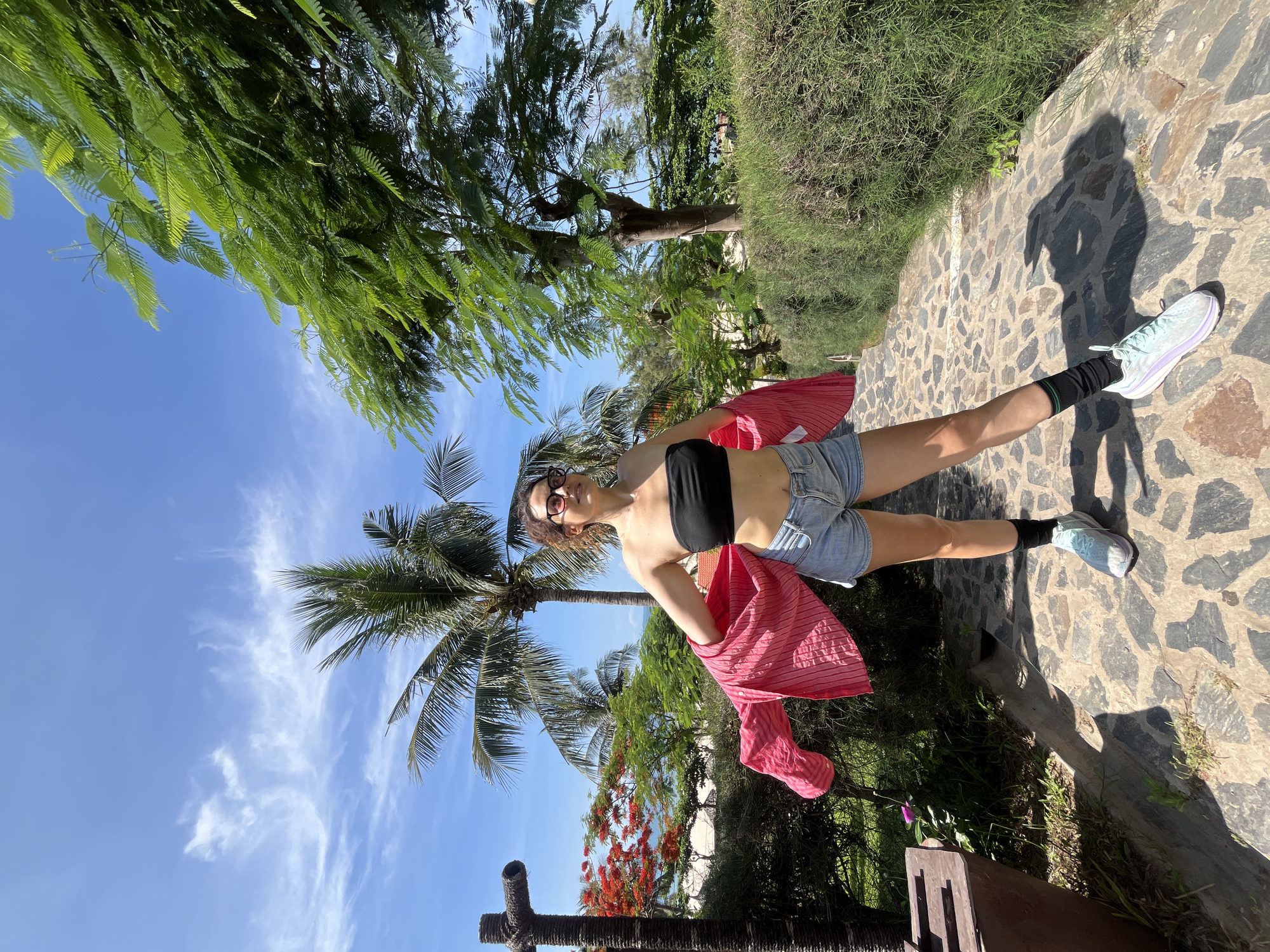 A woman stands on a stone pathway in Phu Thuy Ward, Vietnam, in a tropical outdoor setting. She is wearing a black bandeau top, denim shorts, a pink striped unbuttoned shirt worn open over her shoulders, black socks, and light blue sneakers. She has brown curly hair and is wearing glasses. The background features lush green trees, palm trees, red flowering bushes, and a glimpse of the ocean and beach. The sky is clear blue with scattered white clouds. A brown wooden structure is visible to her left. The pathway is made of irregular grey stones.