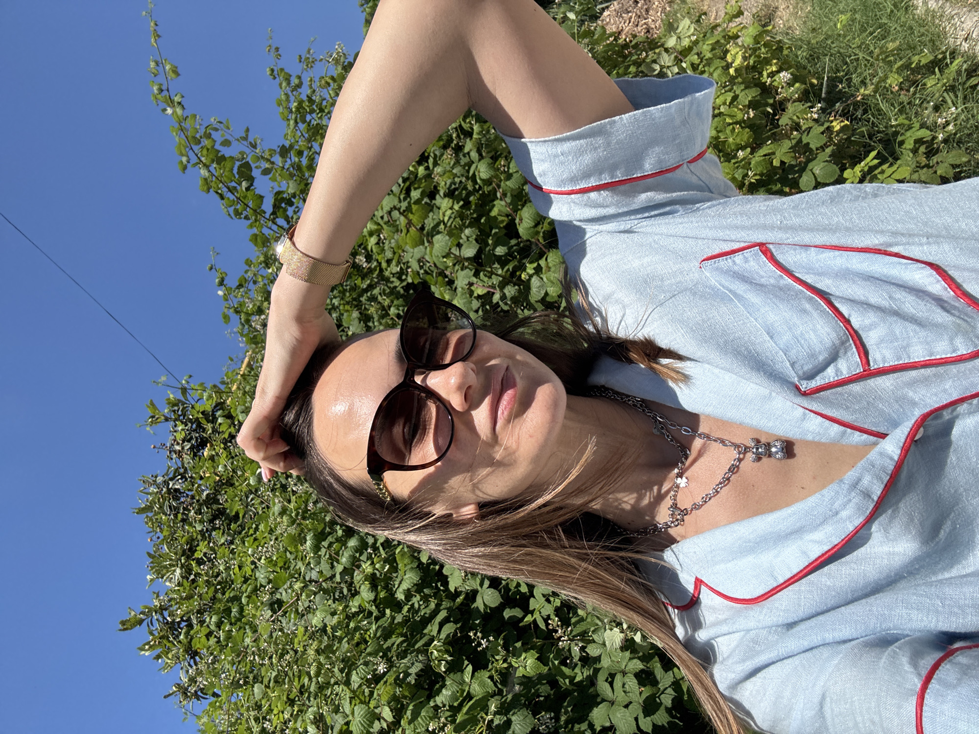 A woman with long brown hair is captured in a selfie outdoors, with a lush green bush and a clear blue sky in the background. She is wearing oversized sunglasses and a light blue collared shirt with red trim. Her right hand is raised, touching her hair. A gold bracelet adorns her left wrist, and a silver necklace with a charm is visible on her chest. The sunlight casts shadows across her face and clothing, indicating a bright, sunny day. The setting appears to be a garden or an area with abundant vegetation, possibly in a suburban or natural environment. There are no indications of Sochi, Russia, within the visual data.