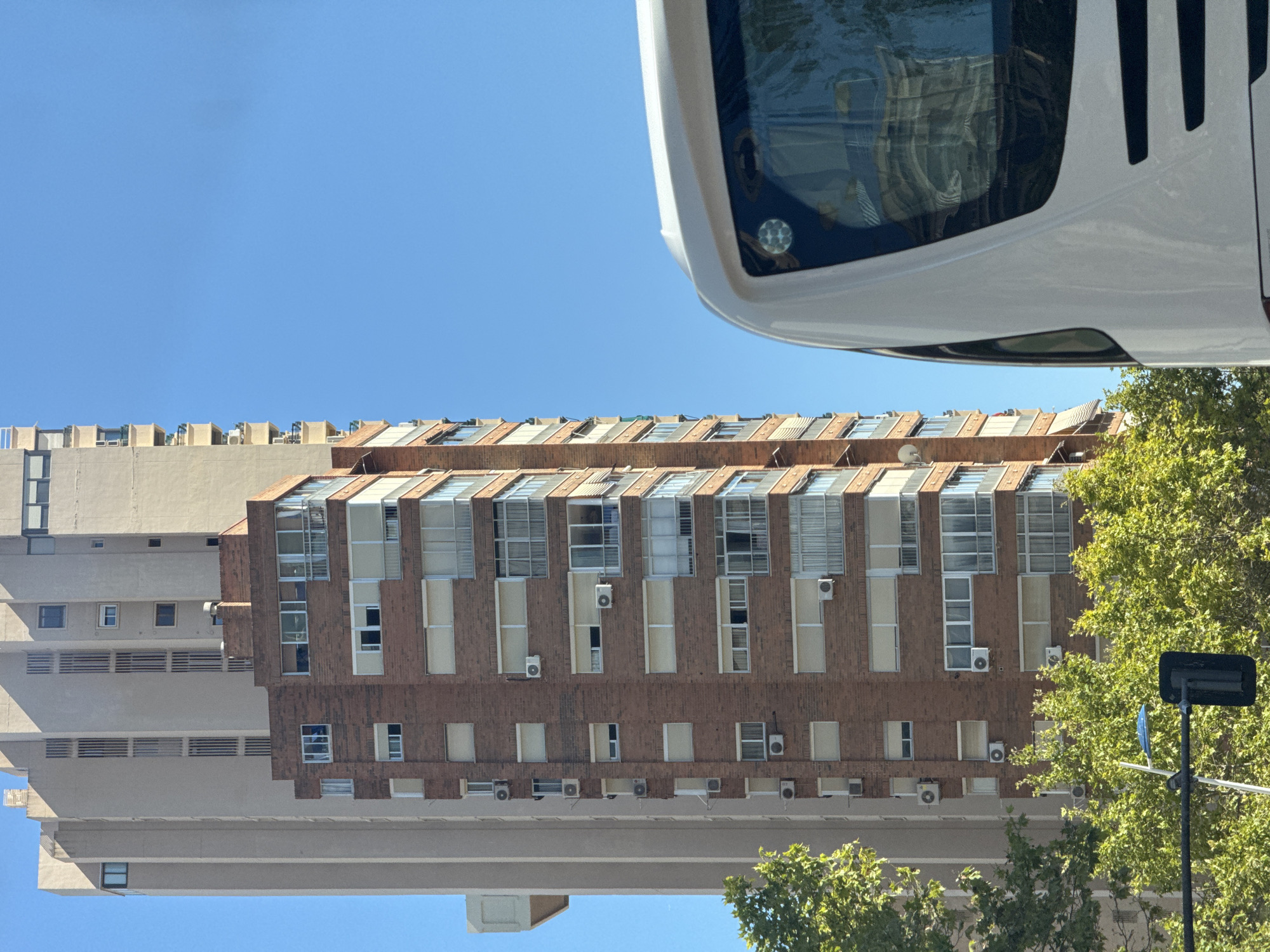 This is a close-up, eye-level, outdoor, daytime photograph taken from a slightly low angle looking up at a bright blue sky. In the foreground, the upper portion of a white bus is visible on the right side of the frame, its windshield reflecting the buildings opposite. Behind the bus, a multi-story brick building with many windows and balconies stands tall, punctuated by several air conditioning units. To the left of the brick building, a taller, lighter-colored building with a more uniform facade is visible. In the lower left of the image, lush green trees partially obscure a street light and traffic signal. The scene appears to be in a dense urban environment, likely Benidorm, Spain, given the architecture typical of the coastal city.