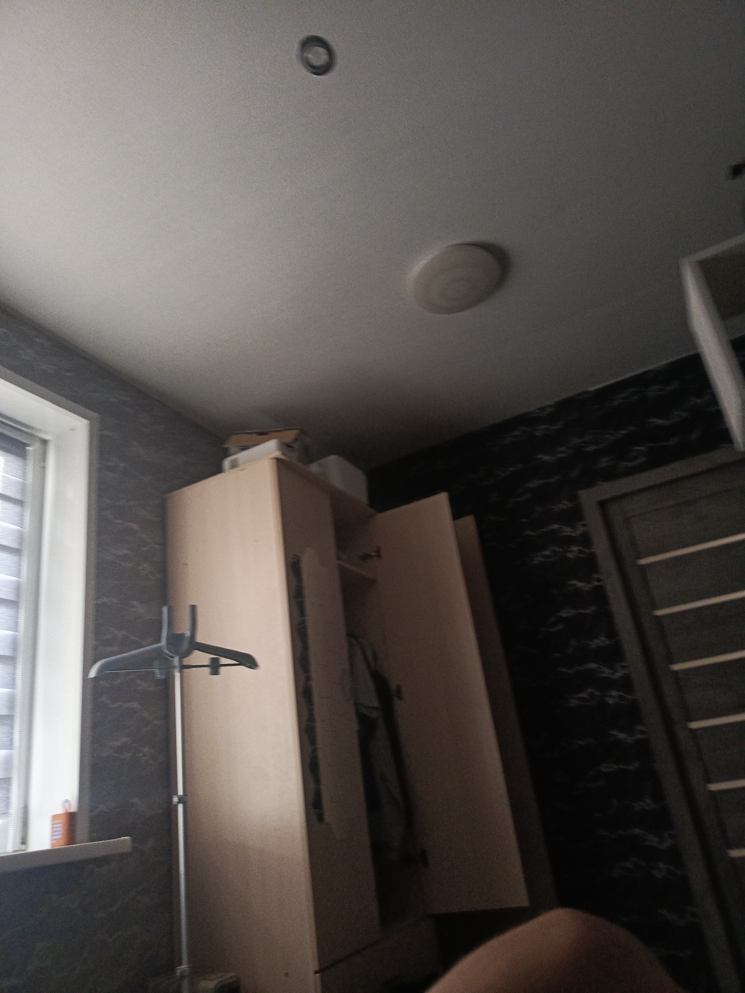 A low-angle, wide shot captures an indoor scene that appears to be a bedroom or closet area in Lesosibirsk, Russia. The dominant elements are a light-colored, wooden wardrobe with its doors ajar, revealing clothes hanging inside. To the left, a window with blinds is visible, and on the windowsill, an orange object, possibly a bottle or container, rests. A clothes hanger on a stand is positioned in front of the window. The walls are covered in dark, patterned wallpaper, and a closed wooden door with horizontal slats is visible on the right side of the frame. The ceiling features two circular light fixtures, one of which is a flush-mounted ceiling light. The overall lighting is dim.