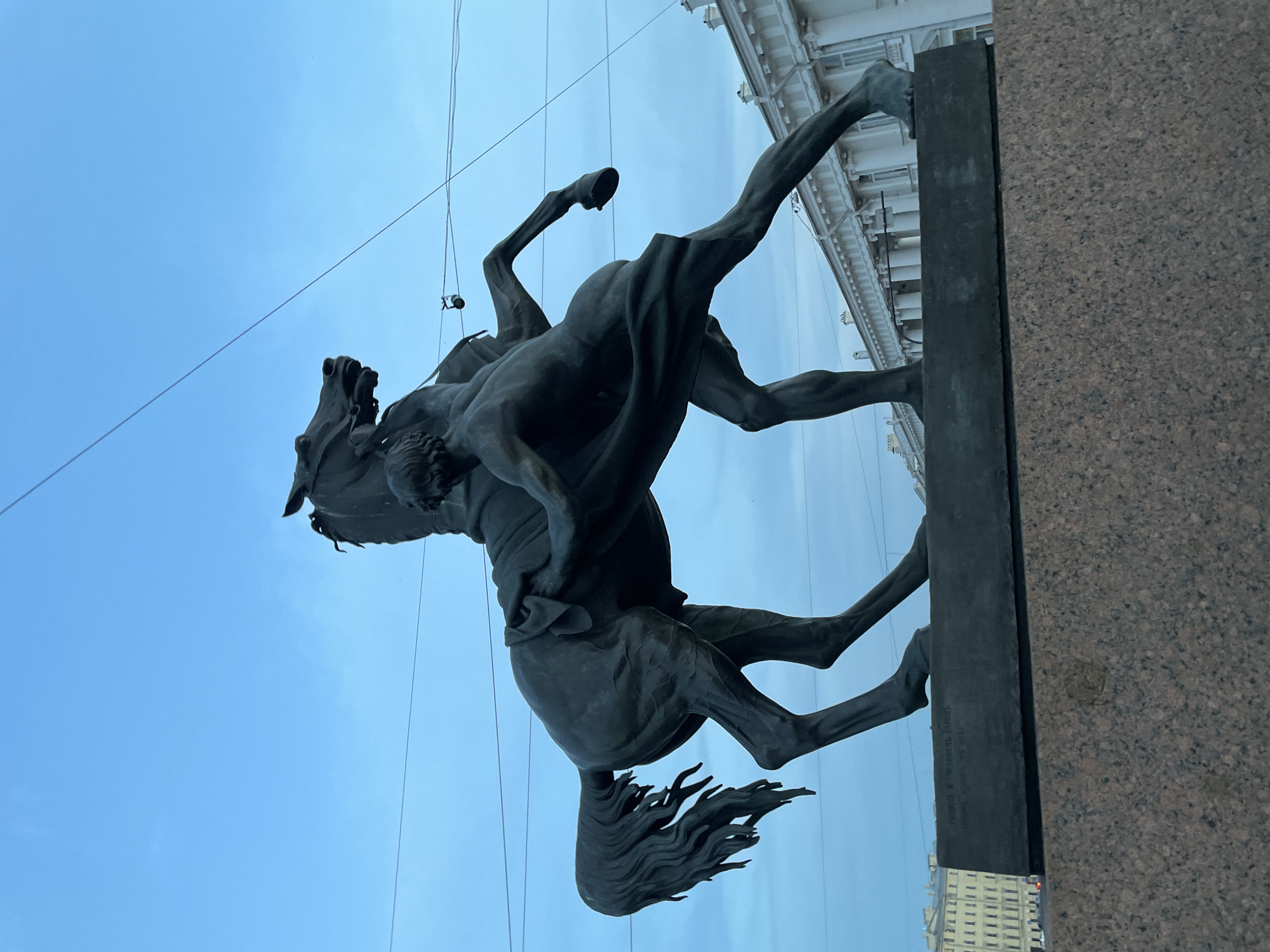The image shows a bronze equestrian statue of a man subduing a horse. The statue is situated on a granite pedestal in the city of Saint Petersburg, Russia. The horse is rearing up on its hind legs, with the man struggling to control it. The man is depicted nude, with his muscular body tense as he pulls on the horse's reins. The statue is dark bronze, contrasting with the clear blue sky behind it. In the background, there are buildings with classical architecture and utility wires crossing the sky. The statue appears to be an outdoor monument, likely in a public square or park. The overall impression is one of strength, struggle, and control.