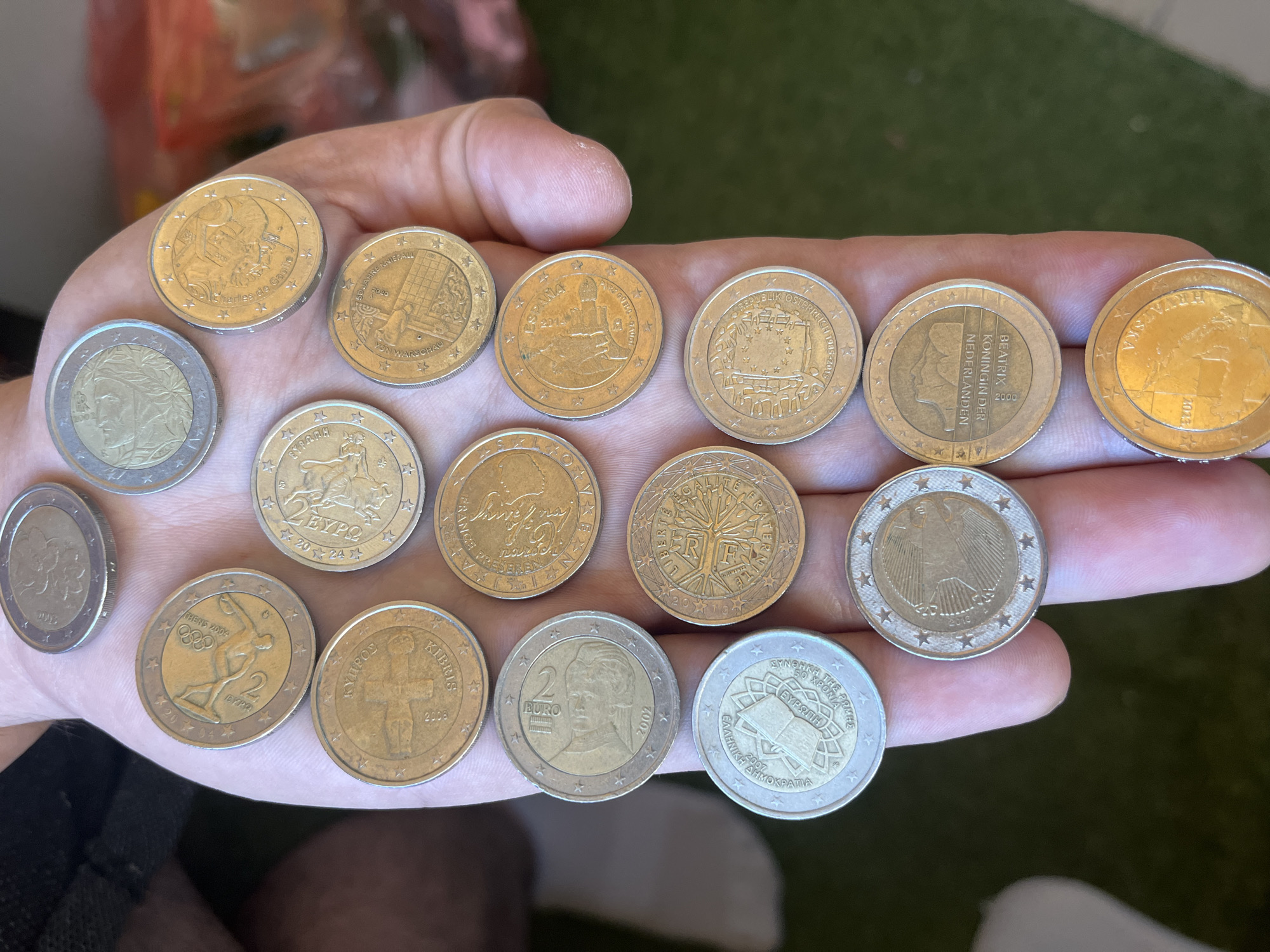 The image shows a human hand holding a collection of various 2 Euro commemorative coins. There are 16 coins visible, each with a unique design. The coins appear to be in circulation and show signs of wear. The coins are displayed on the palm of the hand, and a small portion of the person's arm and torso are visible. The background includes blurred elements such as a white wall and a green surface, possibly a garden. The scene is set in an indoor location. The specific location in Plovdiv, Bulgaria is not indicated in the image.