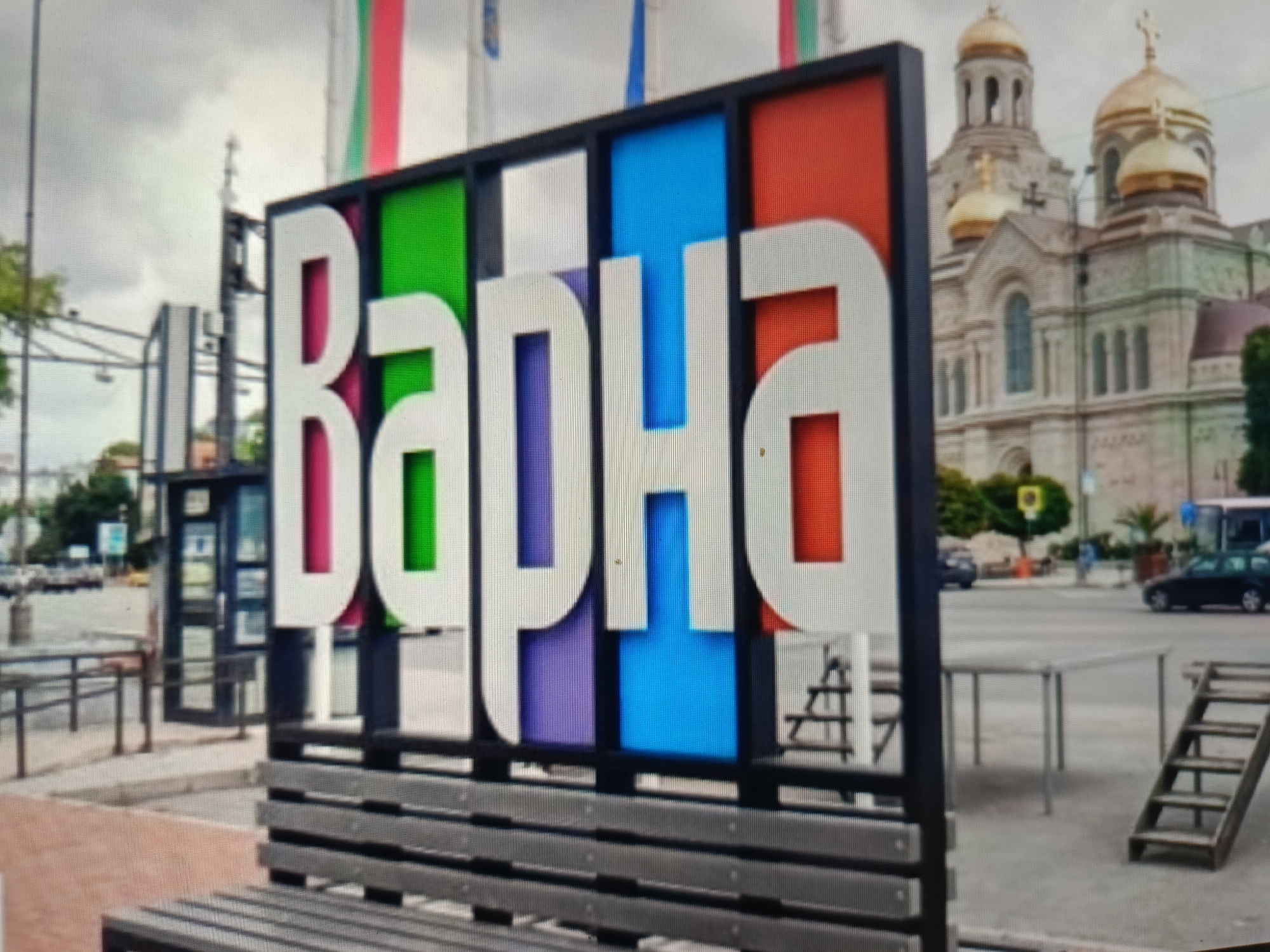 The image depicts a street scene in Varna, Bulgaria, featuring a large, colorful sign that reads "Варна" (Varna). The sign is constructed with large white letters against a backdrop of vertical colored panels, including green, magenta, blue, and red. In the background, a prominent church with golden domes is visible, likely the Dormition of the Mother of God Cathedral, a landmark in Varna. The foreground shows a wooden bench, suggesting a public area or a seating spot near the sign. The environment appears to be an urban setting with streets, sidewalks, and other buildings visible. The sky is overcast.
