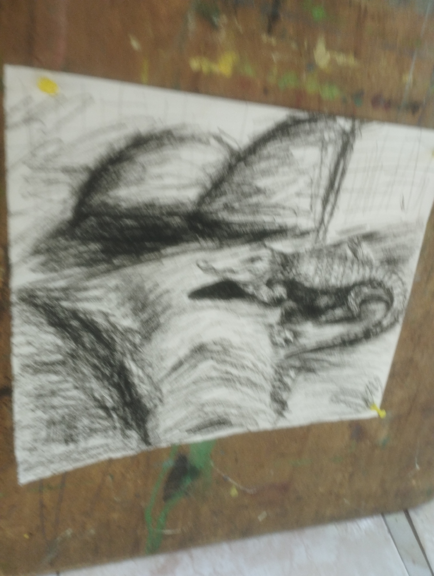 A charcoal sketch of a dragon or mythical creature is displayed on a white sheet of paper, which is attached to a wooden easel. The sketch depicts the creature in a coiled position, with its head resting on the ground. The creature's scales are rendered with rough, energetic charcoal strokes, suggesting texture and depth. The background of the sketch is left mostly blank, with some loosely drawn lines that could represent a landscape or water. The easel itself is weathered and appears to be made of wood, with some smudges of paint visible on its surface. The overall impression is that of an artistic work in progress, with the charcoal sketch being the focal point of the image.
