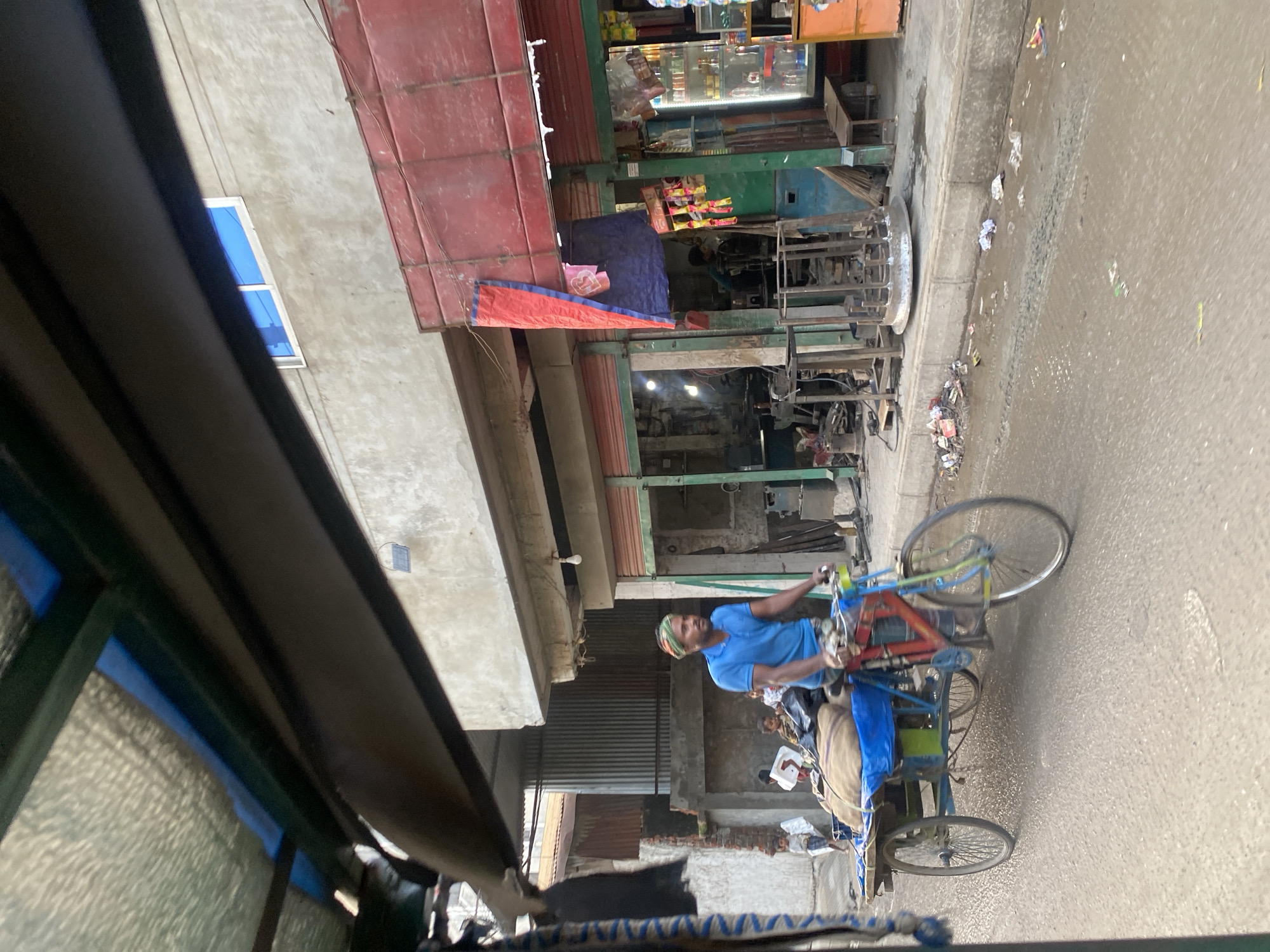 A street in Dhaka, Bangladesh, is depicted from the perspective of a rickshaw, revealing a man driving a cycle rickshaw. The man wears a blue shirt and a colorful head covering. He is pulling a rickshaw laden with goods covered in blue tarps. The road surface is gray and appears somewhat worn. Buildings line the street. The shops have red and green awnings. Some appear to be metalwork shops. There are some people walking on the street.