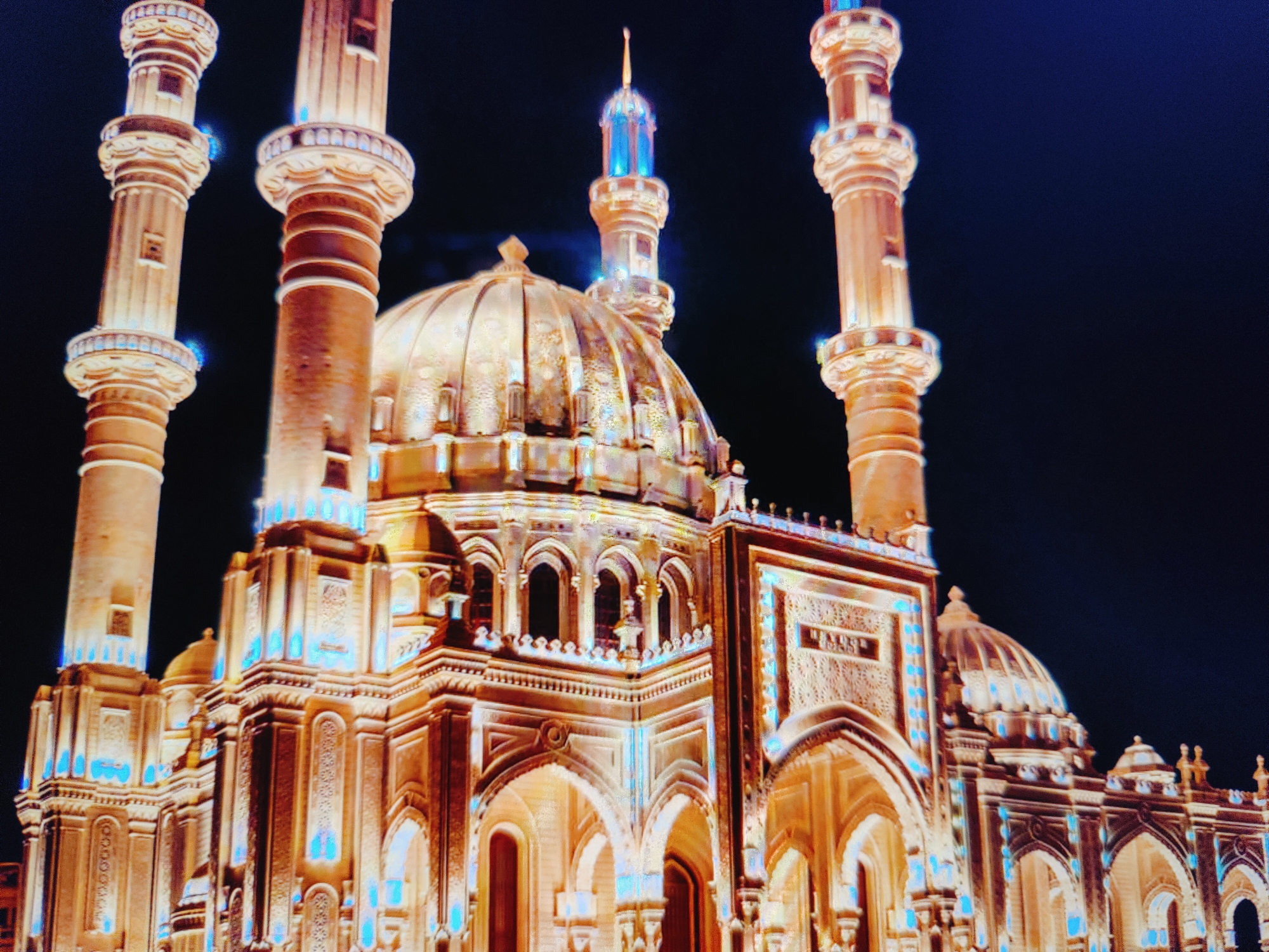 The image shows a night view of the Heydar Aliyev Mosque in Baku, Azerbaijan. The mosque is illuminated with warm, golden lights, highlighting its architectural details. Four tall, slender minarets, characteristic of Islamic architecture, flank the central dome. The dome itself is ornate and rises majestically. Intricate carvings and decorative elements are visible on the mosque's facade, with accents of blue light adding a striking contrast to the golden illumination. The surrounding environment is dark, indicating it is nighttime, with the deep blue sky providing a backdrop. No people are visible in the frame.