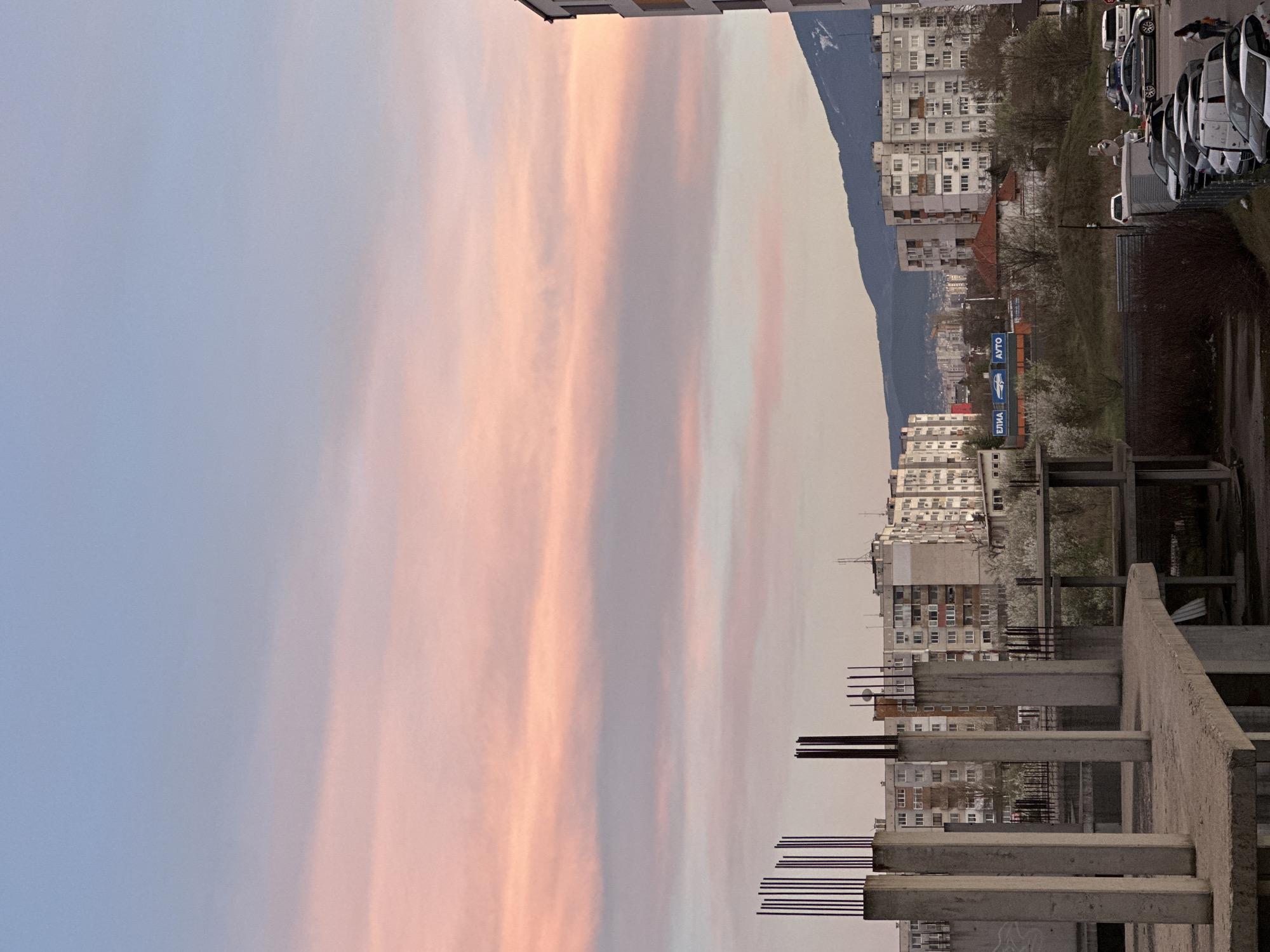 The image depicts a cityscape at twilight, likely in Sofia, Bulgaria, as indicated by the presence of cyrillic script on signage. The foreground shows a concrete structure, possibly an unfinished building, with exposed rebar extending upwards. Beyond this, a dense urban landscape unfolds, consisting of multiple apartment buildings. A mountain range is visible in the background. The sky is dominated by elongated clouds tinted with hues of pink and orange, suggesting a sunset or sunrise. A row of parked cars is visible to the right side of the image. There appears to be at least one person standing near the cars. Vegetation is seen intermixed with the buildings, and a few bare trees and flowering trees.