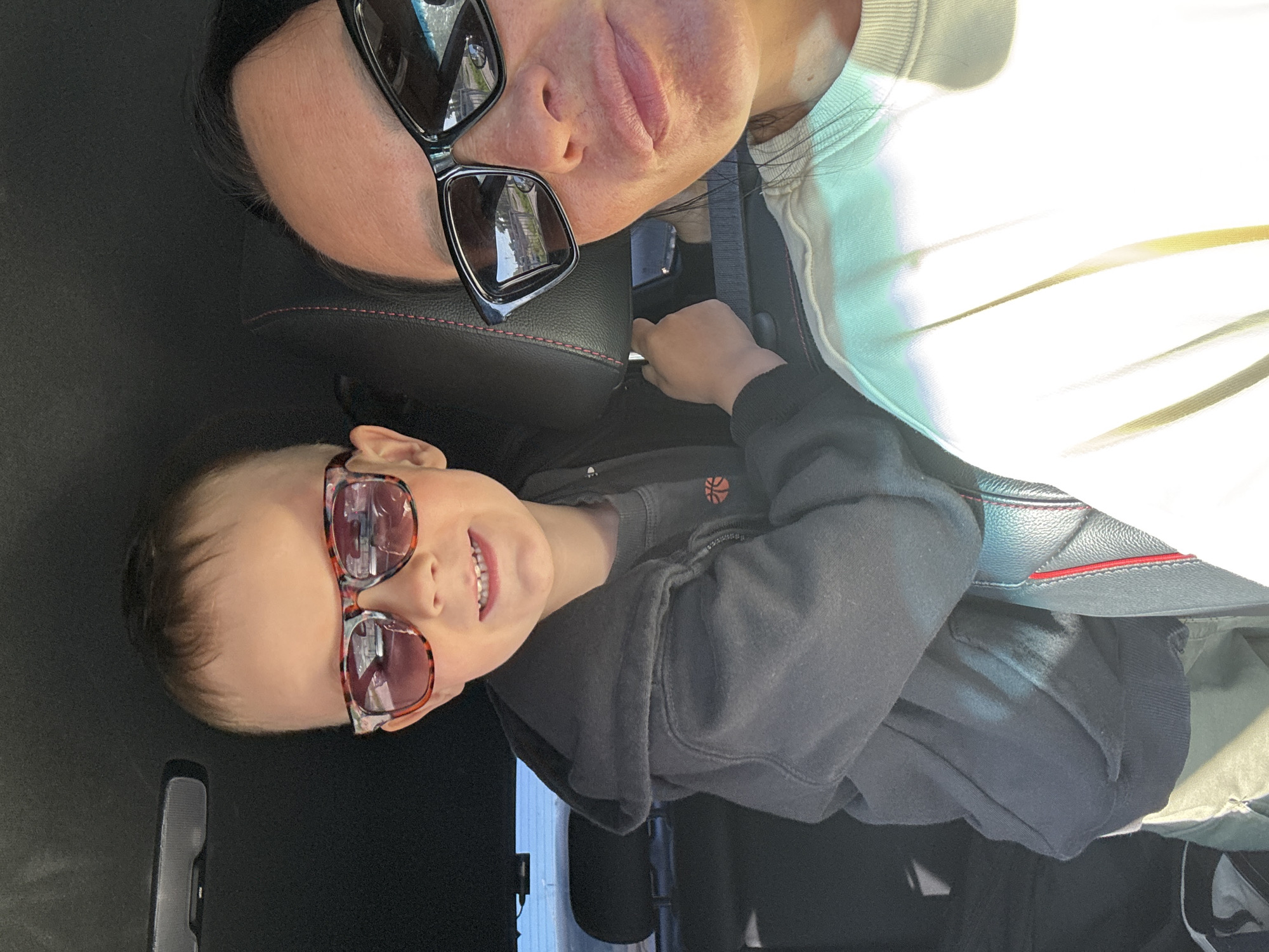 A medium shot shows a woman and a child in a car. The woman, wearing black-rimmed sunglasses, is leaning her head back against the headrest of the car seat. The child, seated beside her, is wearing tortoise-shell pattern sunglasses and a gray hooded sweatshirt with a basketball design. The interior of the car is visible, including the seats, seatbelts, and a rearview mirror. The image provides no definitive information to locate the scene within Chaikovsky, Russia, as no landmarks or features are visible that would indicate a specific location.