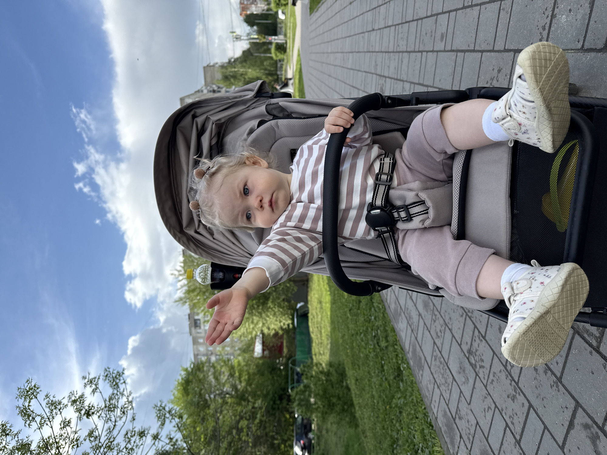 A small child sits in a stroller on a paved walkway. The child is wearing a striped long-sleeved shirt, mauve-colored pants, white socks, and white shoes with floral patterns. A black stroller harness secures the child in the gray stroller. The child is reaching out with their left hand. The background includes green grass, trees, buildings, and a cloudy blue sky. The location is Vladimir, Russia.
