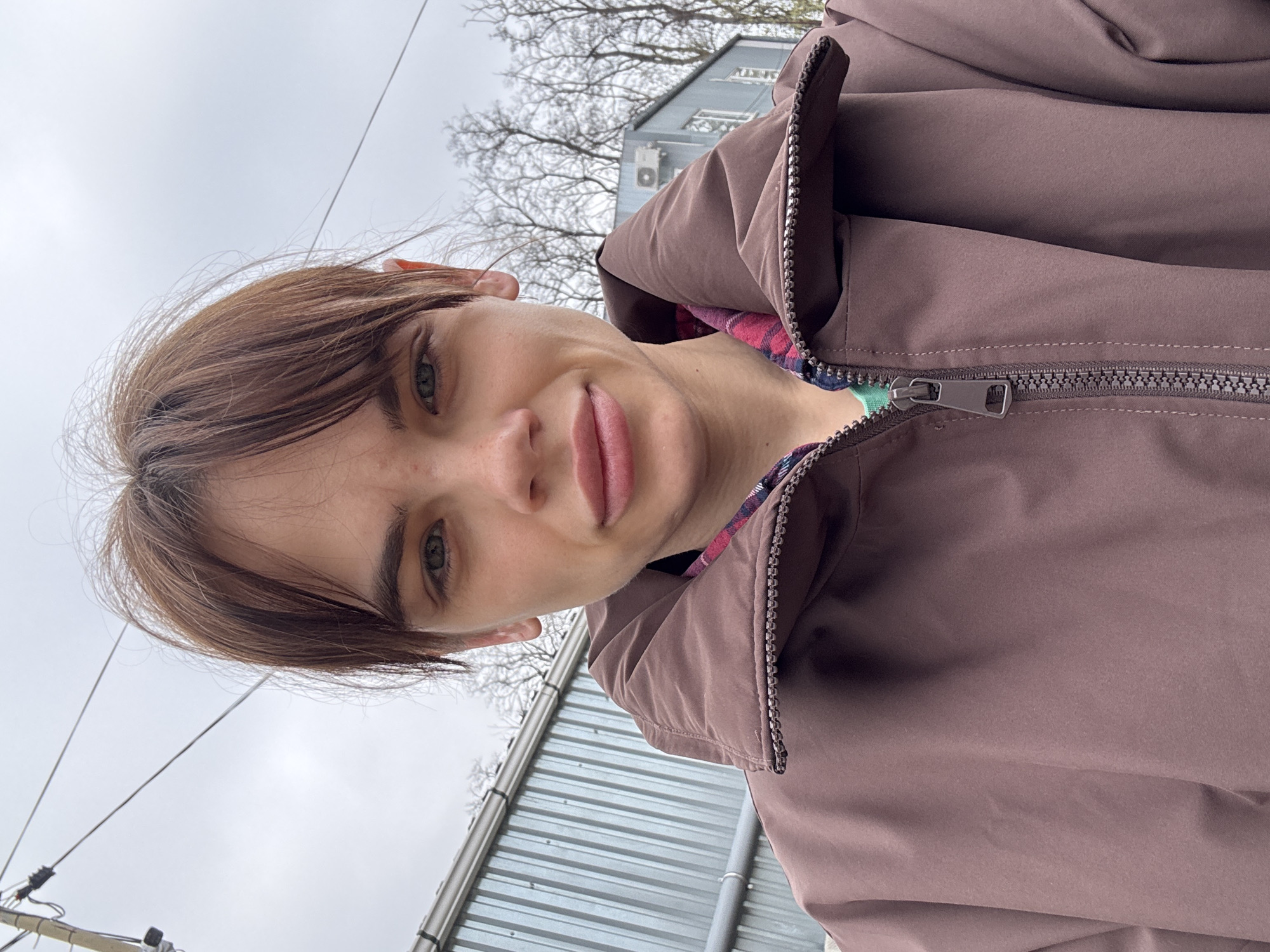 A portrait of a woman with short brown hair and light eyes, smiling at the camera. She is wearing a brown jacket with a plaid lining visible around the collar. The background includes a cloudy sky, bare trees, and a light-colored building. In the lower portion of the image, corrugated metal is visible. While the description asked for the location to be included if it was Rostov-on-Don, Russia, there is no clear indication of the location in the image.