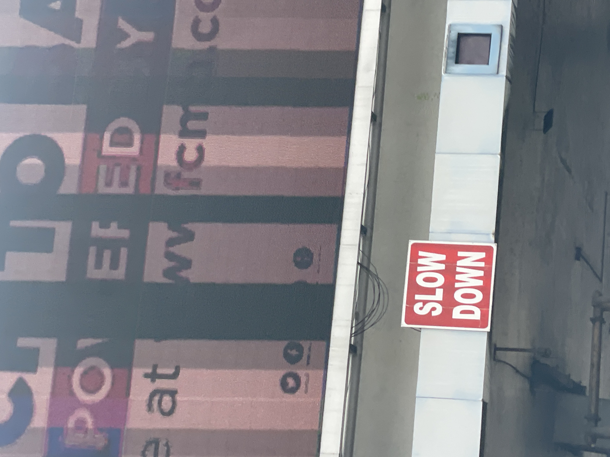 The image shows a vertical view of a section of a building facade. On the left is a large advertisement with text and graphic elements, including the partial URL "www.fcm.co." To the right of the advertisement, a vertical white structure features a "SLOW DOWN" sign with red lettering. The background includes architectural details and an indication of an urban environment. The location, Igboefon, Nigeria is not discernible from the image.