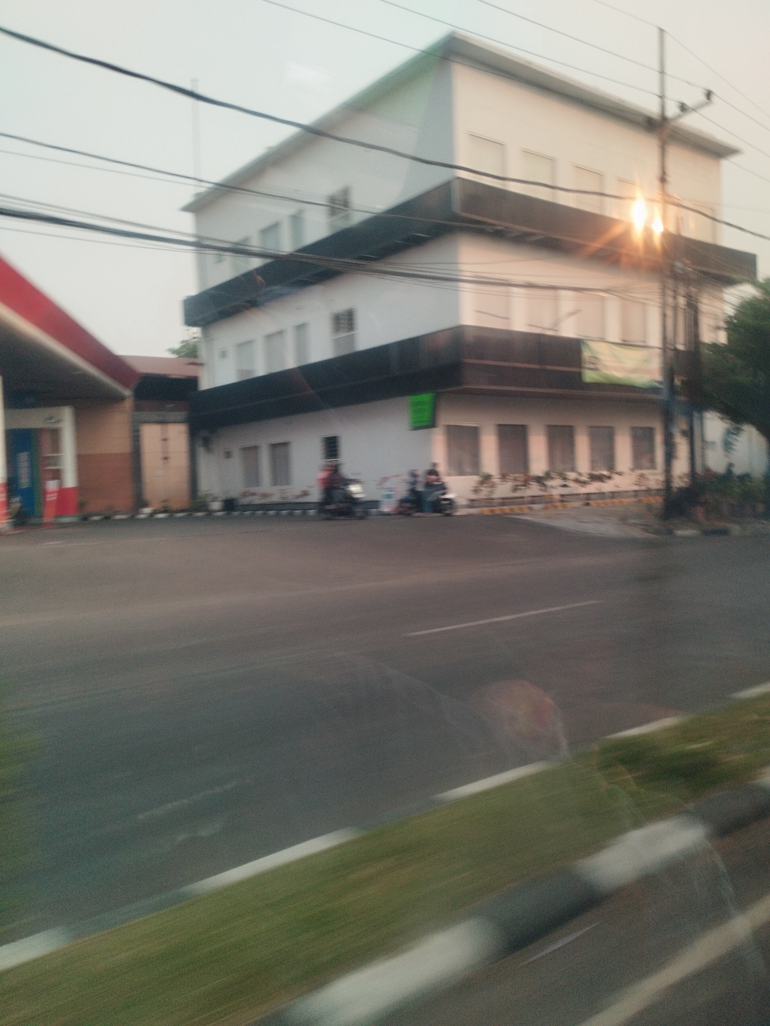 The image depicts a blurred street scene in Surabaya, Indonesia, likely captured from a moving vehicle. In the background, a multi-story white building with dark horizontal accents dominates the frame. The building has numerous windows and appears to be a commercial or office space. A gas station with a red and white awning is partially visible on the left side of the image. Several power lines crisscross the sky above the building and gas station.
In the foreground, a paved road is visible with a grassy median strip bordered by white lines. The motion blur suggests movement, and the lighting indicates it is either early morning or late afternoon. A few motorcycles and figures are present on the road and sidewalk in front of the building, but they are indistinct due to the blur. One individual is riding a motorcycle on the right side of the road. A green banner with some text is hanging on the building. The overall impression is a typical urban street environment in Surabaya.