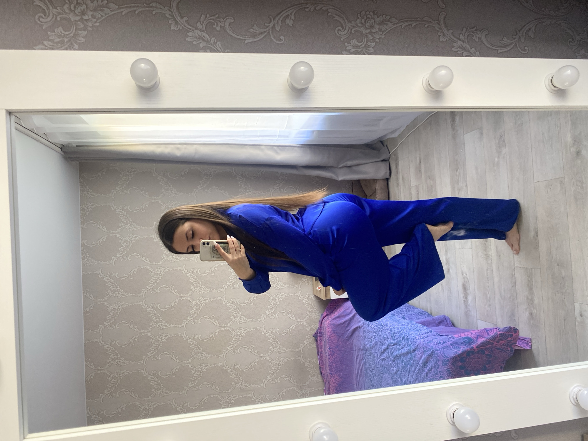 A woman wearing a royal blue jumpsuit is taking a selfie in a mirror with light bulbs attached to the top. The mirror is reflecting a bedroom with light patterned wallpaper, a purple and blue patterned duvet on a bed, and light wooden flooring. The location appears to be in an interior setting. The image doesn't contain any information about the city of Chaikovsky, Russia, which is outside the scope of the scene being depicted.