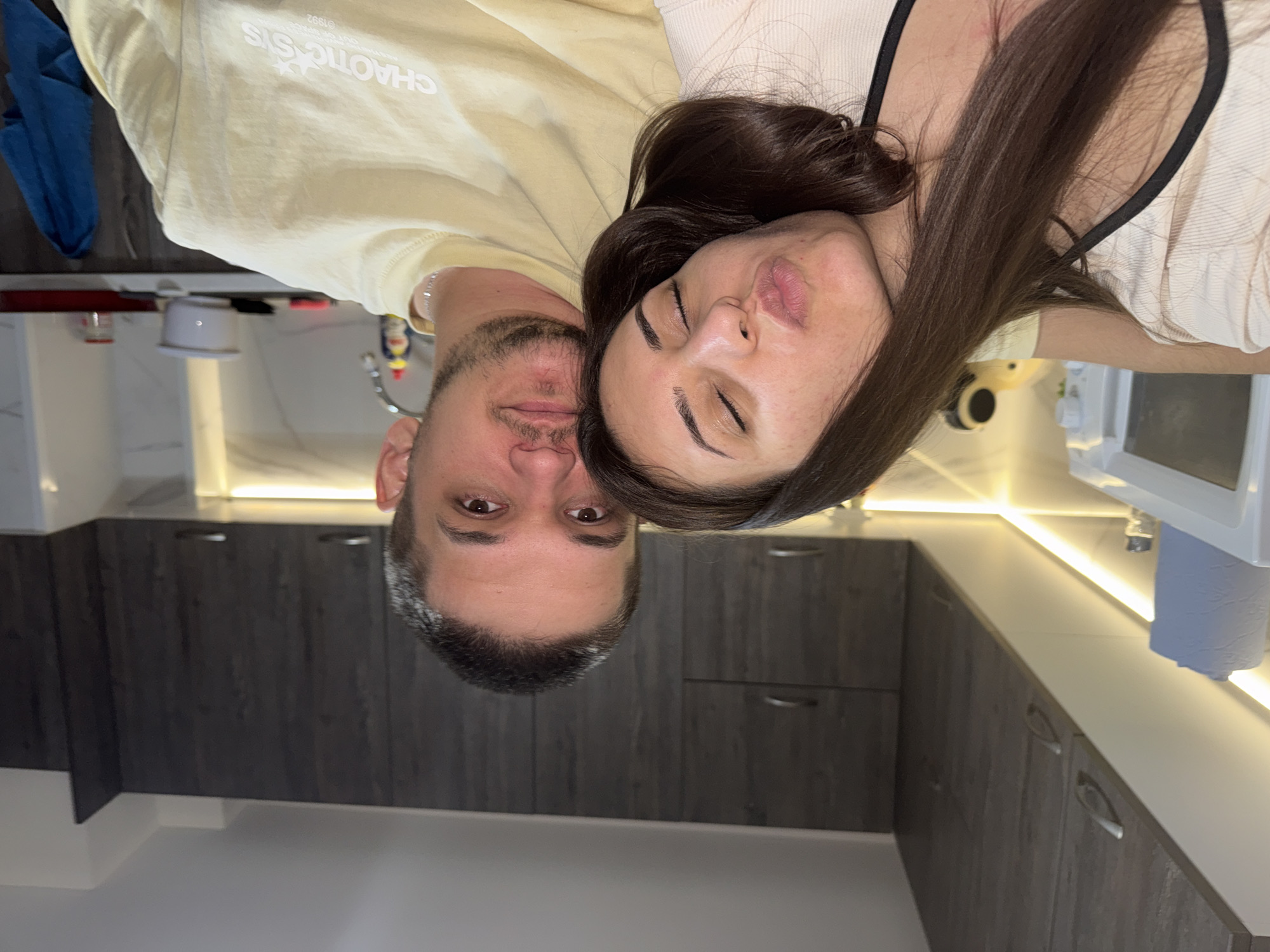The image displays two individuals, a female and a male, situated in what appears to be a modern residential kitchen. The female, positioned on the left, has dark brown hair, closed eyes, and her lips pursed as she leans her head on the male's right shoulder. Her left arm is visible, draped over the male's shoulder. She wears a light-colored top with black strap details. The male, on the right, has short dark hair and light facial stubble, looking directly forward. He wears a light yellow t-shirt with "CHAOTIC SYS" printed on the front. His left arm is around the female.
The background consists of dark wood-grain upper kitchen cabinets with silver handles. Below the cabinets, horizontal strip lighting illuminates a white backsplash and countertop. Visible objects include a white microwave oven with a grey paper towel roll atop it, a small salt shaker, and a yellow toy or decorative item to the far left. On the right side of the male, part of a kitchen sink with a metal faucet and a red-capped bottle of dish soap is visible. A white bowl, a red tray, and a clear jar are on the counter further right. A blue cloth towel hangs on a lower cabinet handle. No specific identifying markers for the city of Plovdiv, Bulgaria, are discernible within the image content.
