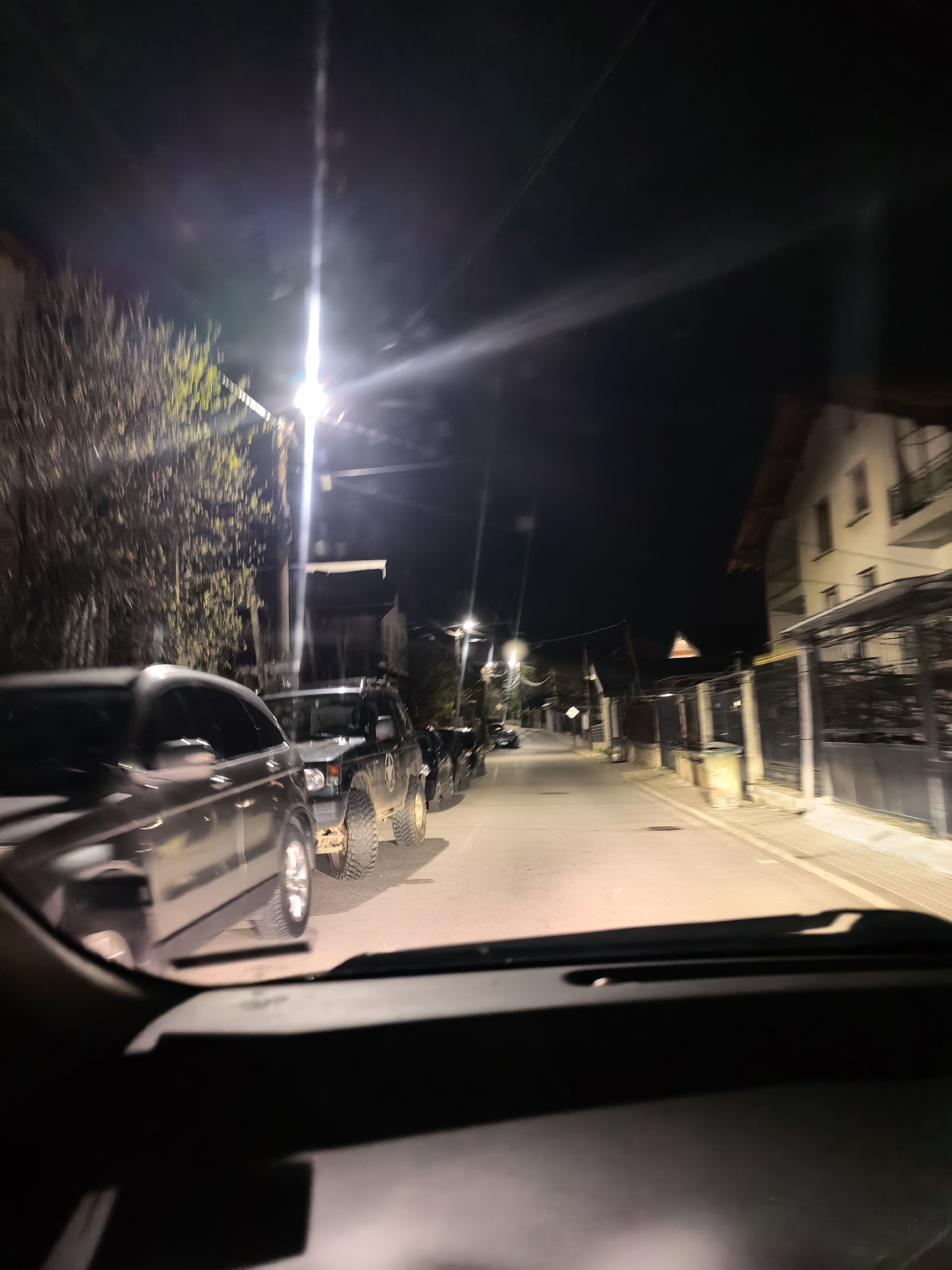 The image shows a street at night, presumably from the perspective of someone sitting in a car. Several cars are parked along the left side of the road. Streetlights illuminate the scene, casting strong light and shadows. Buildings and trees line the street. The exact location is unknown.