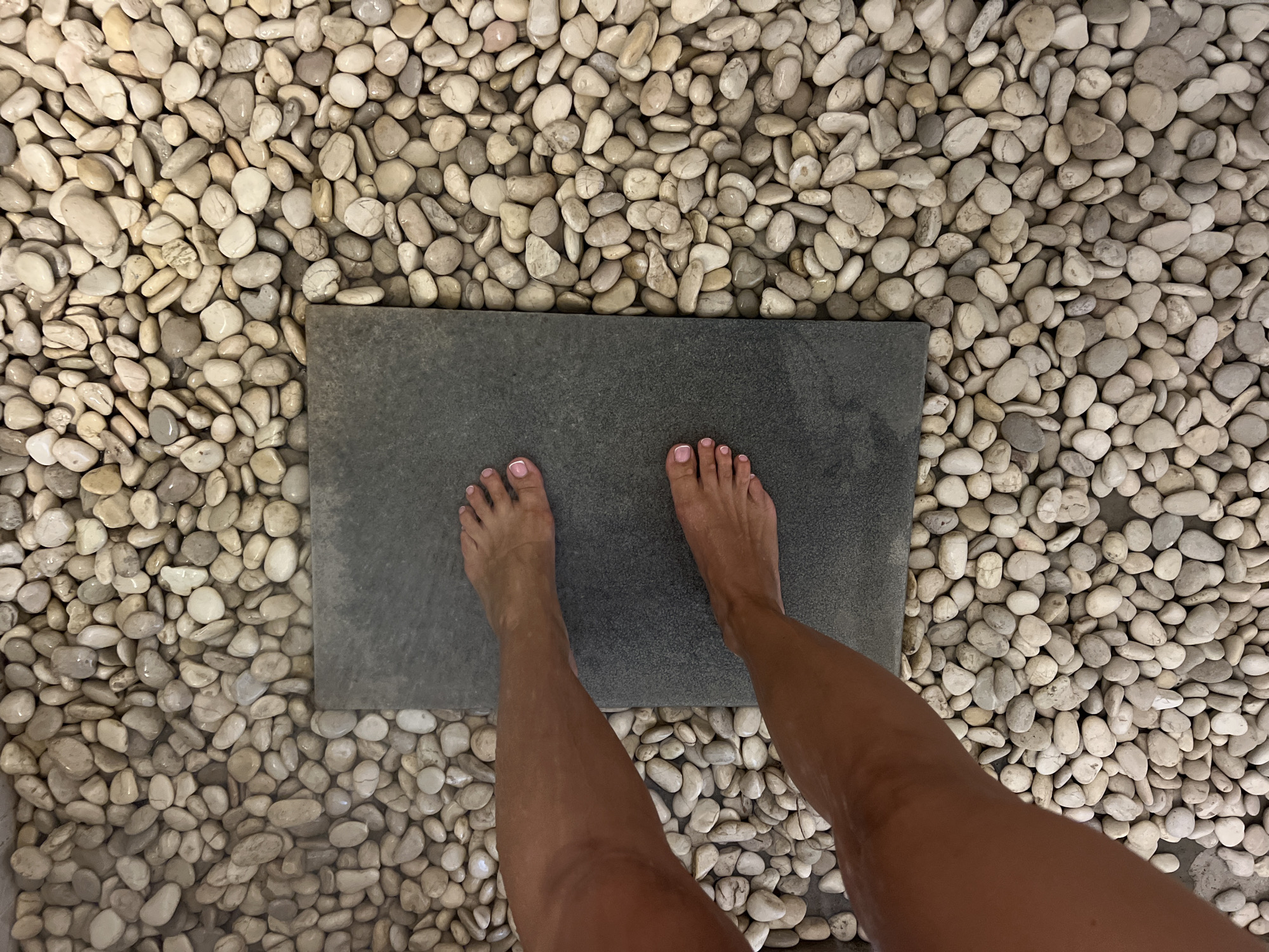 The image shows a person's legs and feet from a top-down perspective. The person is standing with their feet on a dark gray, rectangular slab. This slab is surrounded by a dense bed of smooth, light-colored pebbles. The pebbles appear to be wet, suggesting a bathroom or spa-like setting, possibly a shower floor or foot bath.
The person's feet are bare and have light pink nail polish on their toenails. Their legs are tanned and appear wet, further reinforcing the idea of a water-related environment. The lighting is bright, indicating daytime, though no direct sunlight or specific weather conditions are visible. The scene is devoid of any visible text.
The overall impression is one of relaxation or a moment of transition, perhaps after a swim or shower, in a location designed for comfort and sensory experience. The combination of the cool, smooth slab and the textured pebbles creates a tactile contrast. The setting in North Kuta, Indonesia, might suggest a tropical or resort-like environment, where such natural materials are commonly used in design.