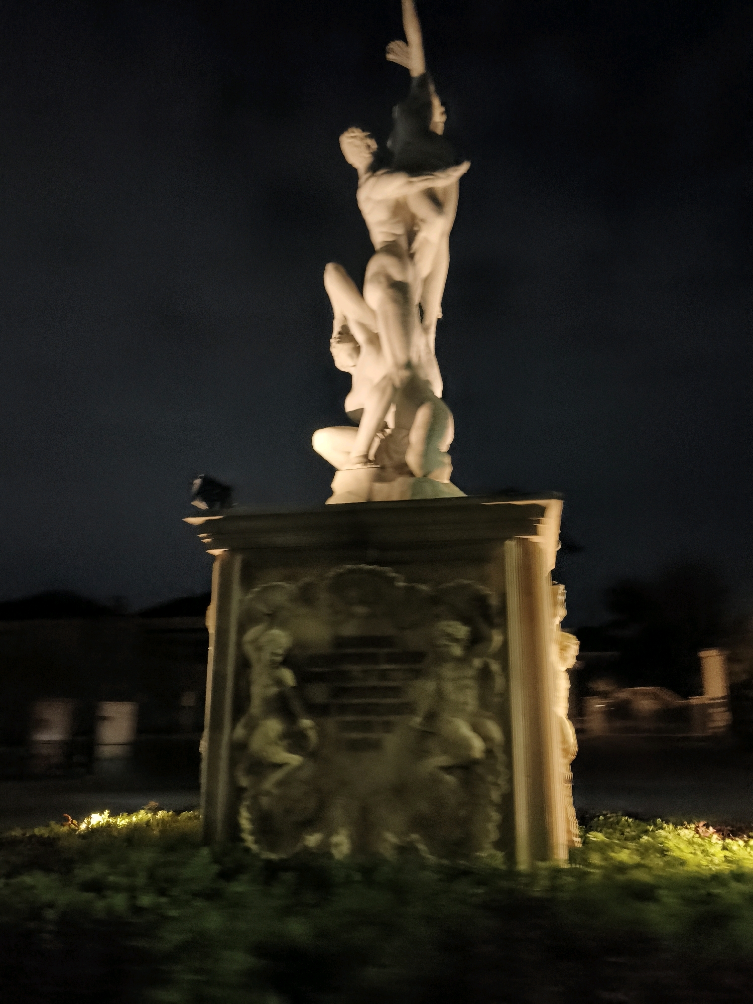 The image shows a blurred, night-time view of a large stone sculpture on a pedestal. The sculpture depicts several nude figures in a dynamic pose, seemingly struggling or lifting. The pedestal has intricate carvings on its sides, featuring human figures and decorative elements. The scene is illuminated by artificial lighting, casting a warm glow on the sculpture and the surrounding vegetation. In the background, blurred structures and trees are visible, suggesting an outdoor urban environment. The overall impression is one of grandeur and artistry, despite the blurriness caused by low light and possibly movement during capture. The sculpture is located at the front of the Grahadi Building, East Java, in the city of Surabaya, Indonesia.