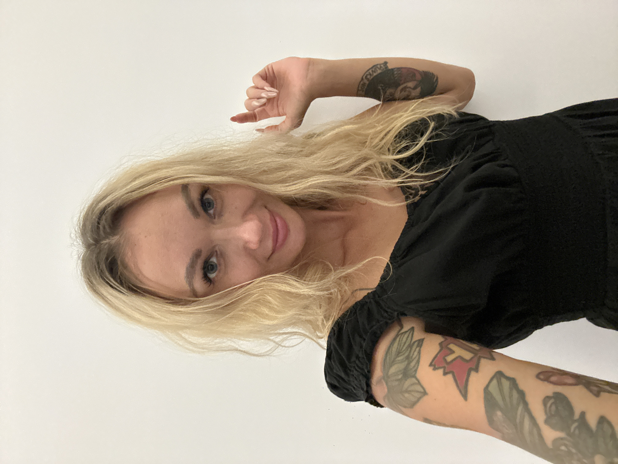 A fair-skinned woman with blonde, shoulder-length hair and blue eyes is depicted in a selfie taken indoors. She is wearing a black dress with gathered sleeves and a fitted bodice. The woman is smiling and looking directly at the camera. She has visible tattoos on both forearms and on the back of her hand. The background is a plain white wall. It is not possible to determine from the image if this was taken in Mũi Né, Vietnam.