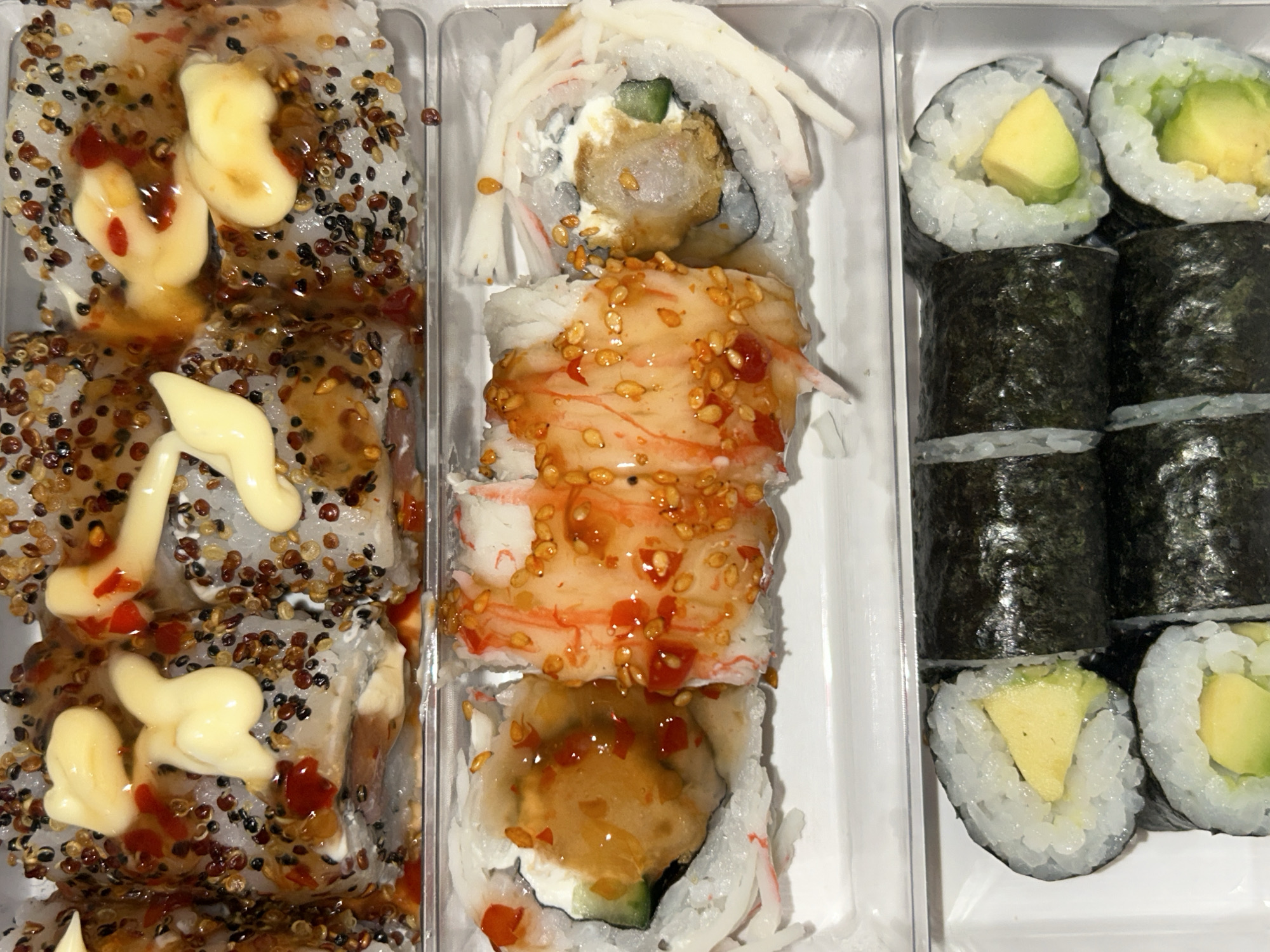 The image shows three plastic trays of sushi. The tray on the left contains sushi rolls coated in sesame seeds, drizzled with a brown sauce and topped with a mayonnaise-like sauce. The middle tray has sushi rolls with white rice on the outside, decorated with shredded crab sticks and a sweet, reddish sauce. The tray on the right contains maki sushi rolls with avocado in the center and nori seaweed wrapping some of them. The location is not discernible from the image.
