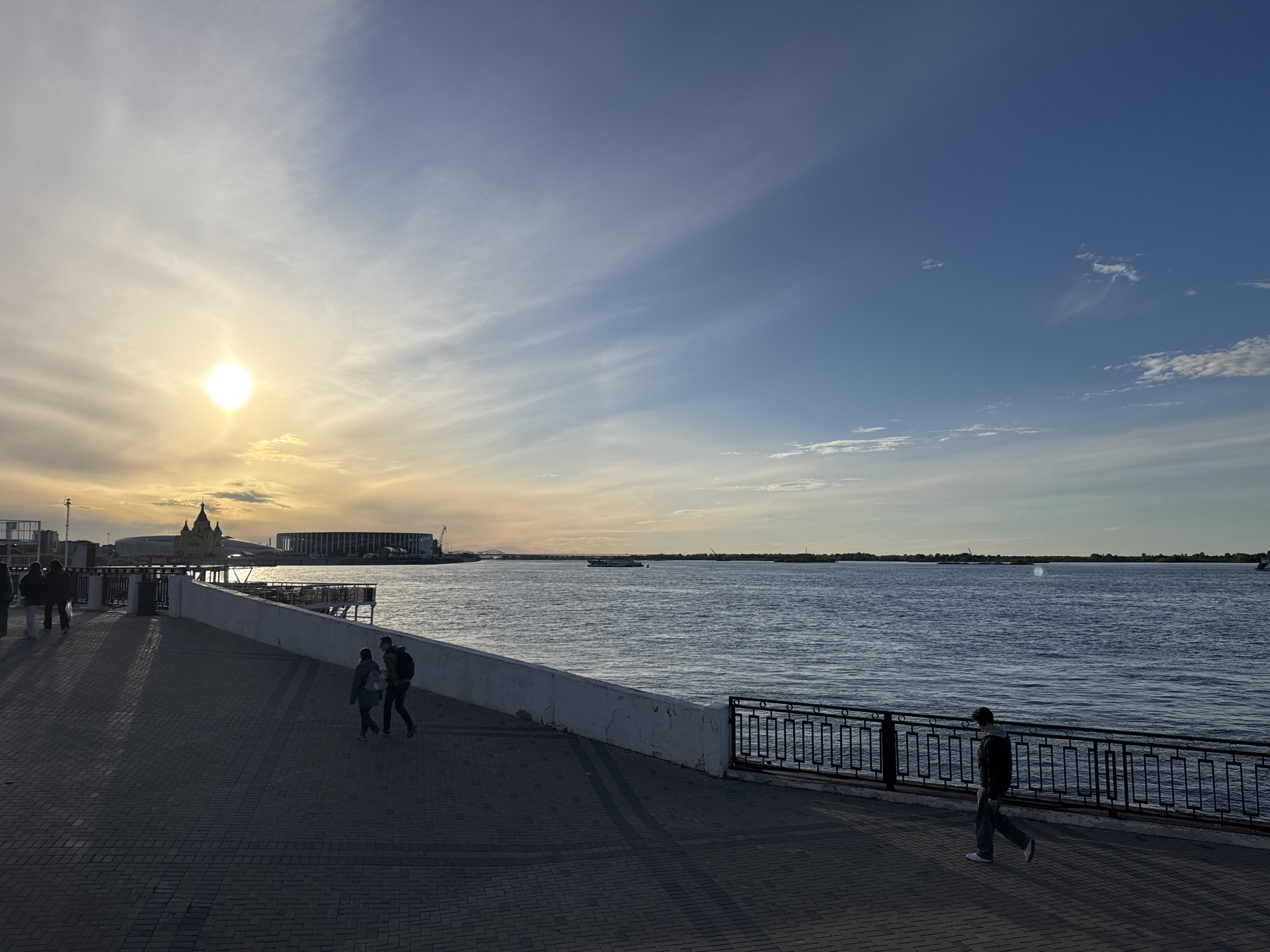The image depicts a waterfront promenade in Nizhny Novgorod, Russia, at dusk or dawn. A brick-paved walkway runs along the river, bordered by a low, white wall on one side and a black metal railing on the other. Several people are visible: a couple walking together near the wall, another person walking solo near the railing, and a small group of people in the distance near a structure with architectural details that appear to be Russian Orthodox. The river is wide and calm with a few small boats visible in the distance. Across the water, the opposite bank is visible with trees and buildings. The sky is a mix of blue and light clouds, with the sun low on the horizon, casting a soft glow on the scene. The overall lighting suggests the possibility of a late afternoon or early morning.