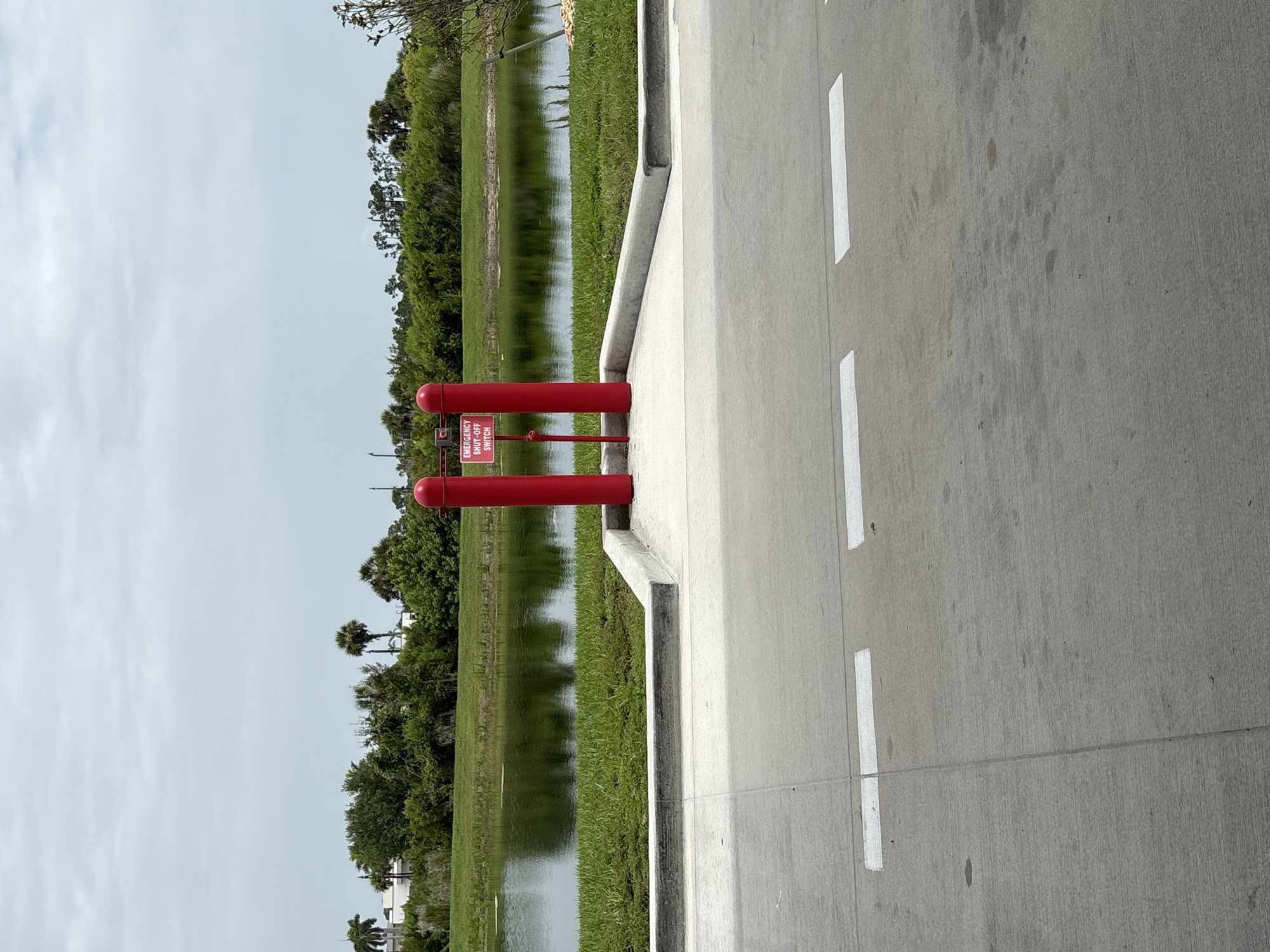 In North Port, United States of America, this image captures a paved area in the foreground with two white rectangular markings on the concrete. Beyond the paved area, a raised concrete structure with a grey curb is visible, leading to two bright red bollards. A red sign reading "EMERGENCY SHUT-OFF SWITCH" is affixed to one of the bollards. In the middle ground, a body of water reflects the overcast sky and is bordered by lush green grass and dense foliage. The background features a line of trees, including palm trees, and some buildings. The overall scene appears to be an outdoor industrial or utility area.