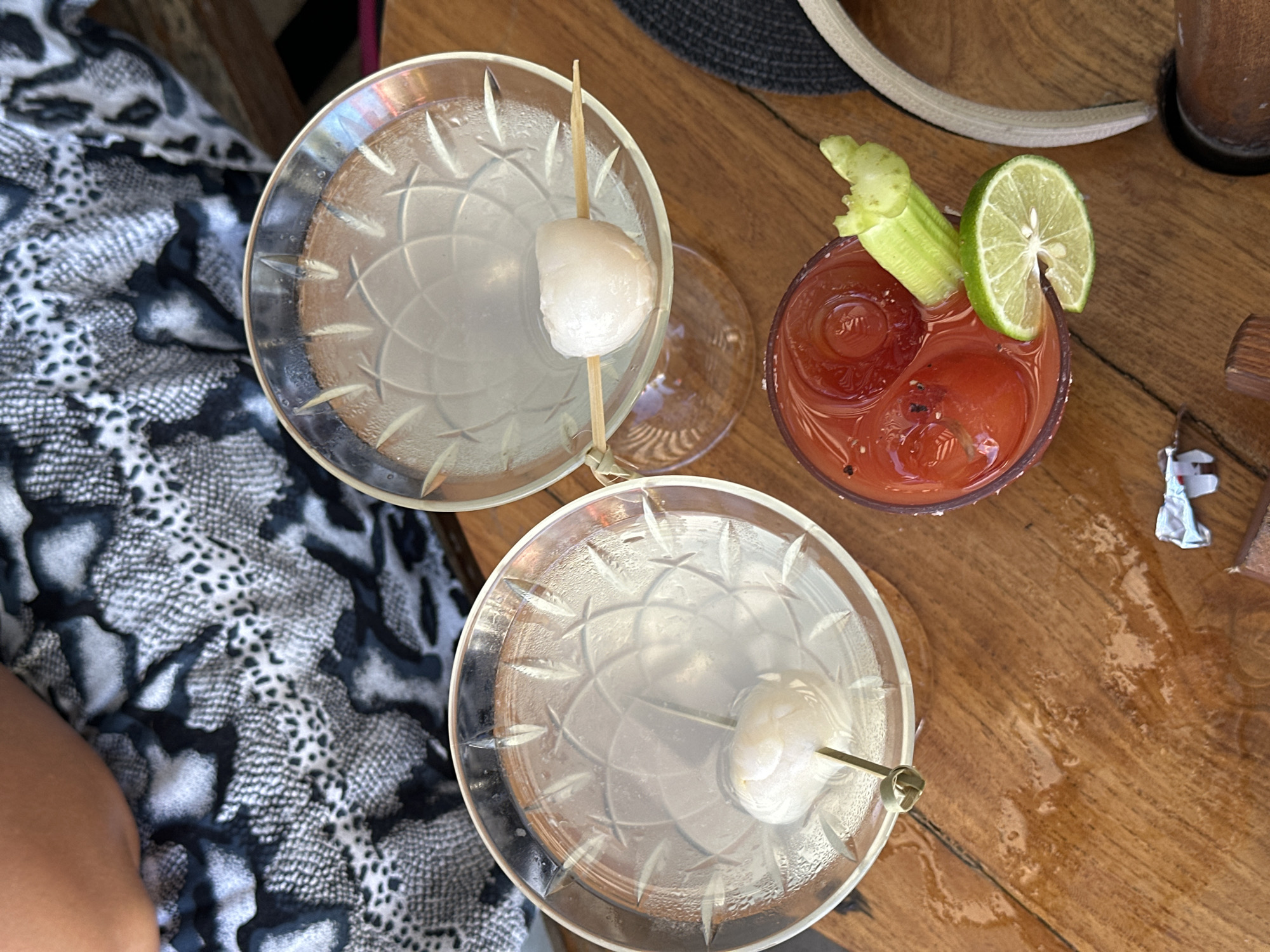 From an overhead perspective, the image showcases a wooden table setting with three drinks: two cocktails in martini glasses, each garnished with a lychee on a skewer, and a Bloody Mary decorated with a celery stalk and a lime wedge. The table's surface exhibits wet spots, possibly from condensation or spills. A portion of a patterned fabric, resembling a snake or animal print, is visible to the left, suggesting someone is seated at the table. A partial view of a woven hat is seen in the upper right corner. The setting could be a restaurant or bar in Kuta, Indonesia, given the style of drinks and the wooden furniture, which aligns with common establishments in the area.