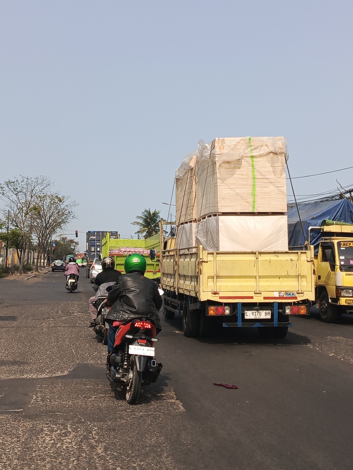 The image depicts a street scene in Surabaya, Indonesia, characterized by moderate traffic under a clear sky. The foreground shows the rear of a red motorcycle with a rider wearing a black leather jacket and a green helmet. The motorcycle's license plate is partially visible, reading "L 2371 DGU". To the right, a yellow flatbed truck is heavily laden with stacks of light-colored wooden boards wrapped in clear plastic and secured with green straps. The truck's license plate is visible as "L 9170 BH". Further back, other vehicles are present, including another truck with a green cargo bed, cars, and more motorcycles. The road surface appears worn, with distinct markings. Trees line the left side of the road, and buildings and power lines are visible in the background, suggesting an urban environment. The overall atmosphere is that of a typical day with ongoing transportation activities.