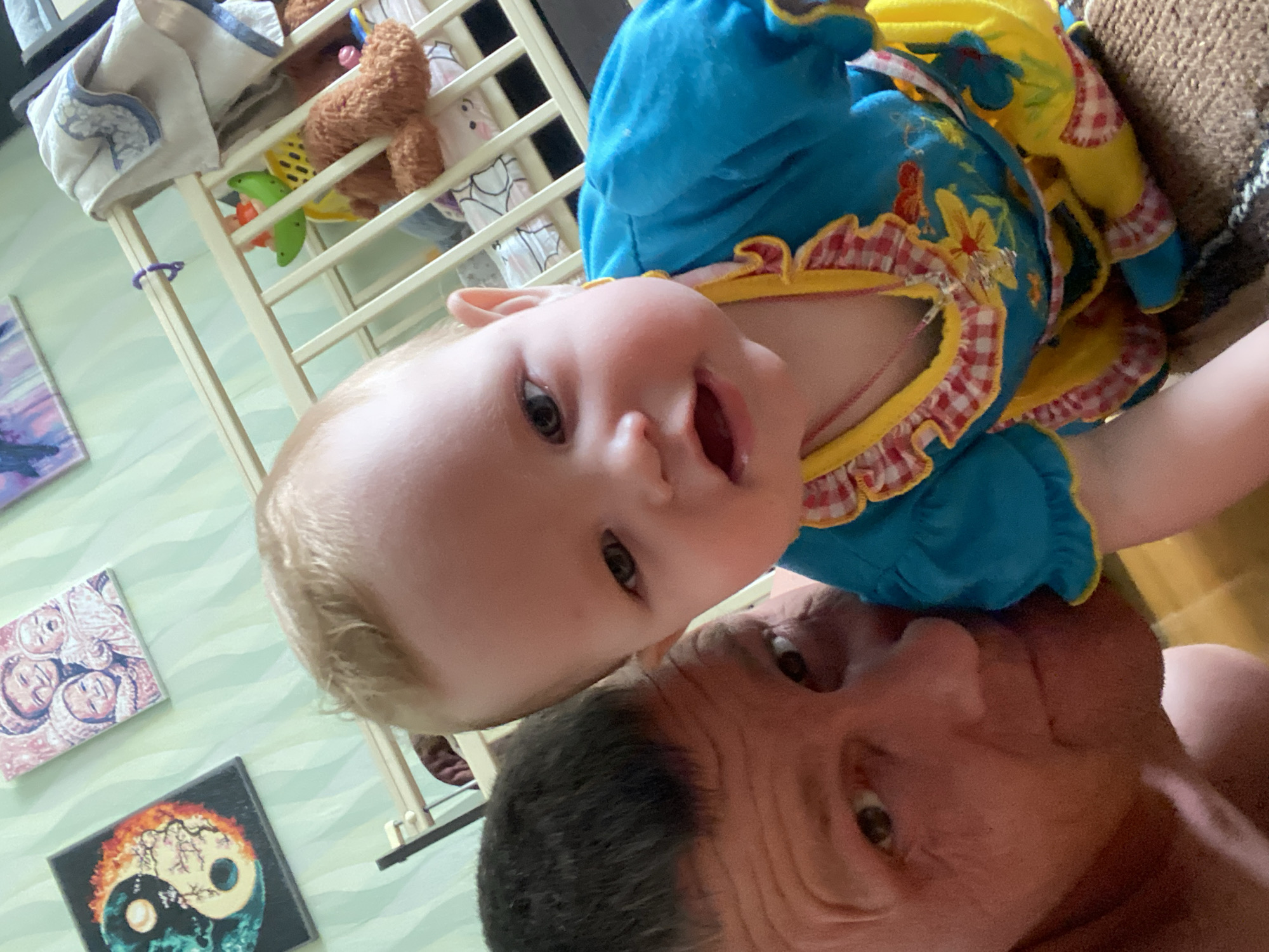A Caucasian baby wearing a blue and yellow dress is being held by an adult Caucasian male with short gray hair. The baby is looking directly at the camera with an open mouth, while the adult is looking up at the baby. The background includes a crib with toys, two framed pictures on a green wall, and a brown upholstered surface. There is no information to indicate that this scene is located in the city of Фоки, Russia.