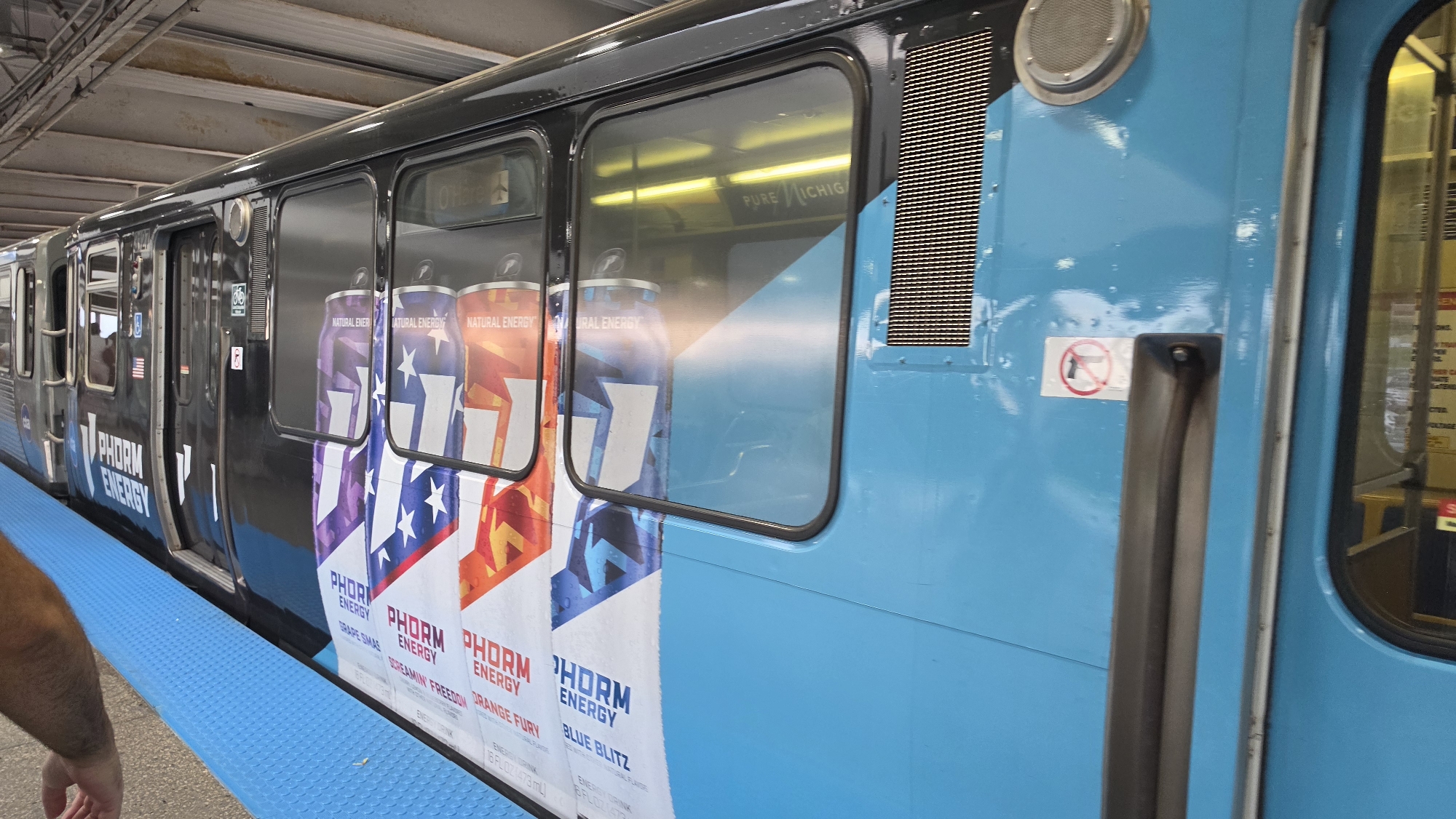 A bright blue and black CTA train is stopped at a station. The train cars are adorned with a colorful advertisement for "PHORM ENERGY" drinks, featuring images of cans with patriotic designs and the text "NATURAL ENERGY". The advertisement wraps around the train, showcasing different flavors such as "GRAPE SMASH", "SCREAMIN' FREEDOM", "ORANGE FURY", and "BLUE BLITZ". The train is positioned alongside a blue platform with a textured surface, commonly found in urban transit environments. In the reflection on the train windows, the word "MICHIGAN" is visible, hinting at a location in or around Chicago, Illinois, which serves the surrounding suburbs including Rosemont, United States of America. The scene is well-lit, suggesting it is daytime.