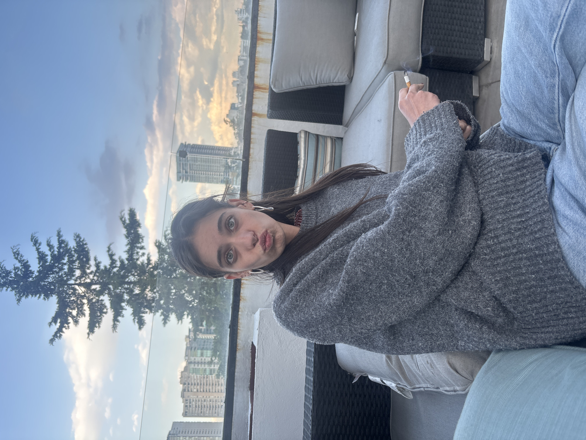 A young woman sits on a patio in Huixquilucan de Degollado, Mexico, holding a lit cigarette in her right hand. She wears a gray sweater and light blue jeans. Behind her is a view of the city skyline, including a tall apartment building. A tree is also visible. She is looking at the camera with pursed lips. The furniture is patio-style wicker with light-colored cushions.