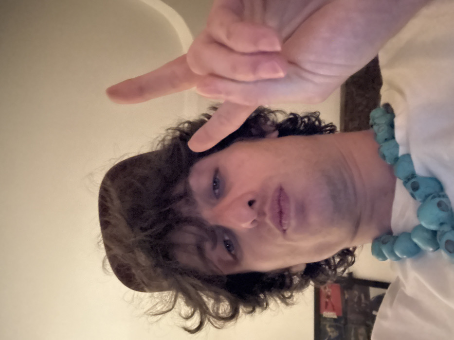 A man with curly hair and a brown hat is facing downward. He wears a white shirt and a necklace of large, round, turquoise beads. He points his index finger up toward the top of the frame. Part of a doorway is visible at the top, and a framed picture can be seen in the background. The image is rotated to the side.